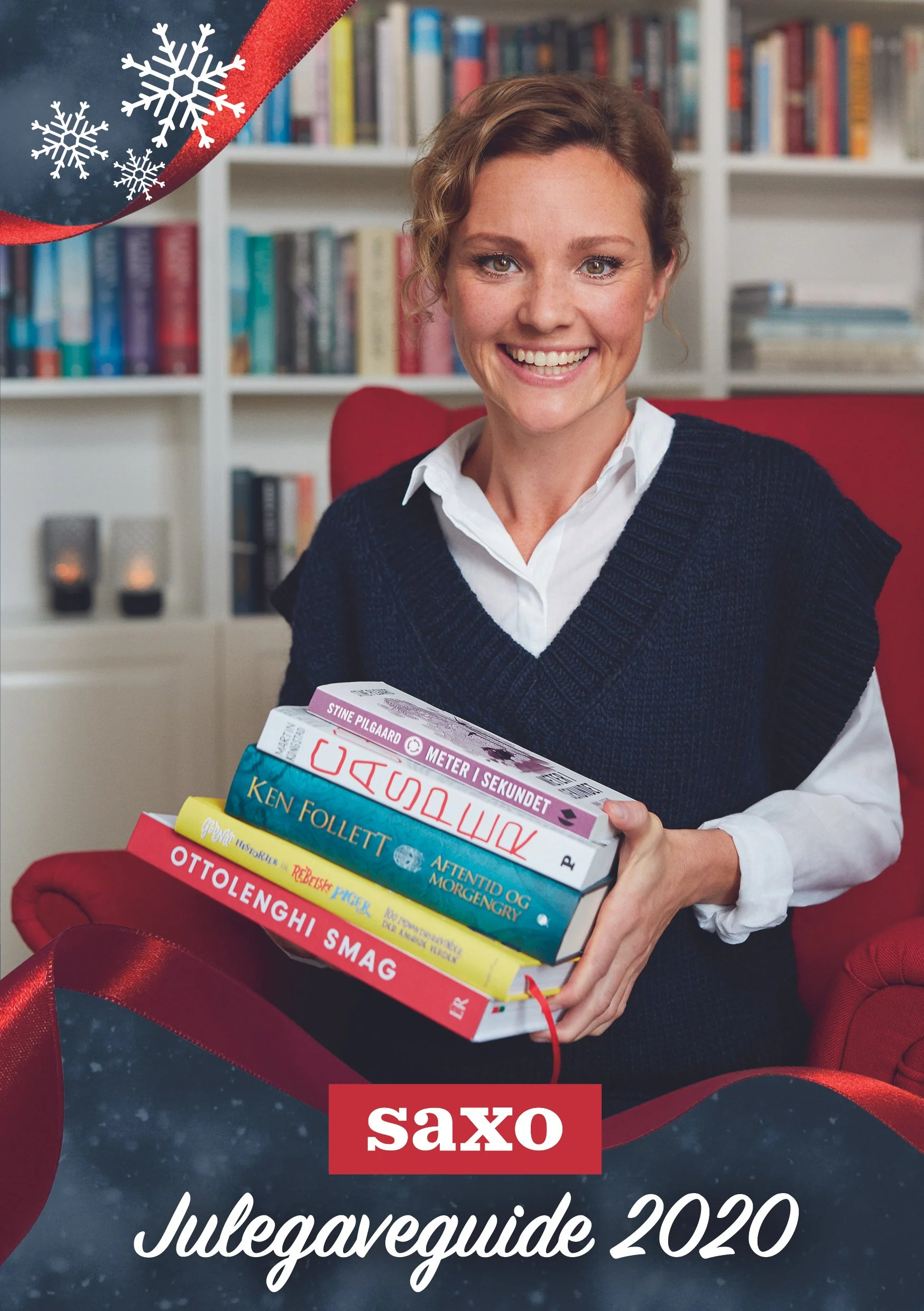 Portrait Lise Rønne for Saxo - A woman with short, curly brown hair and a white shirt under a dark knitted vest smiles while holding several books in her hands. Behind her is a bookshelf with various colorful books in a well-lit room.
