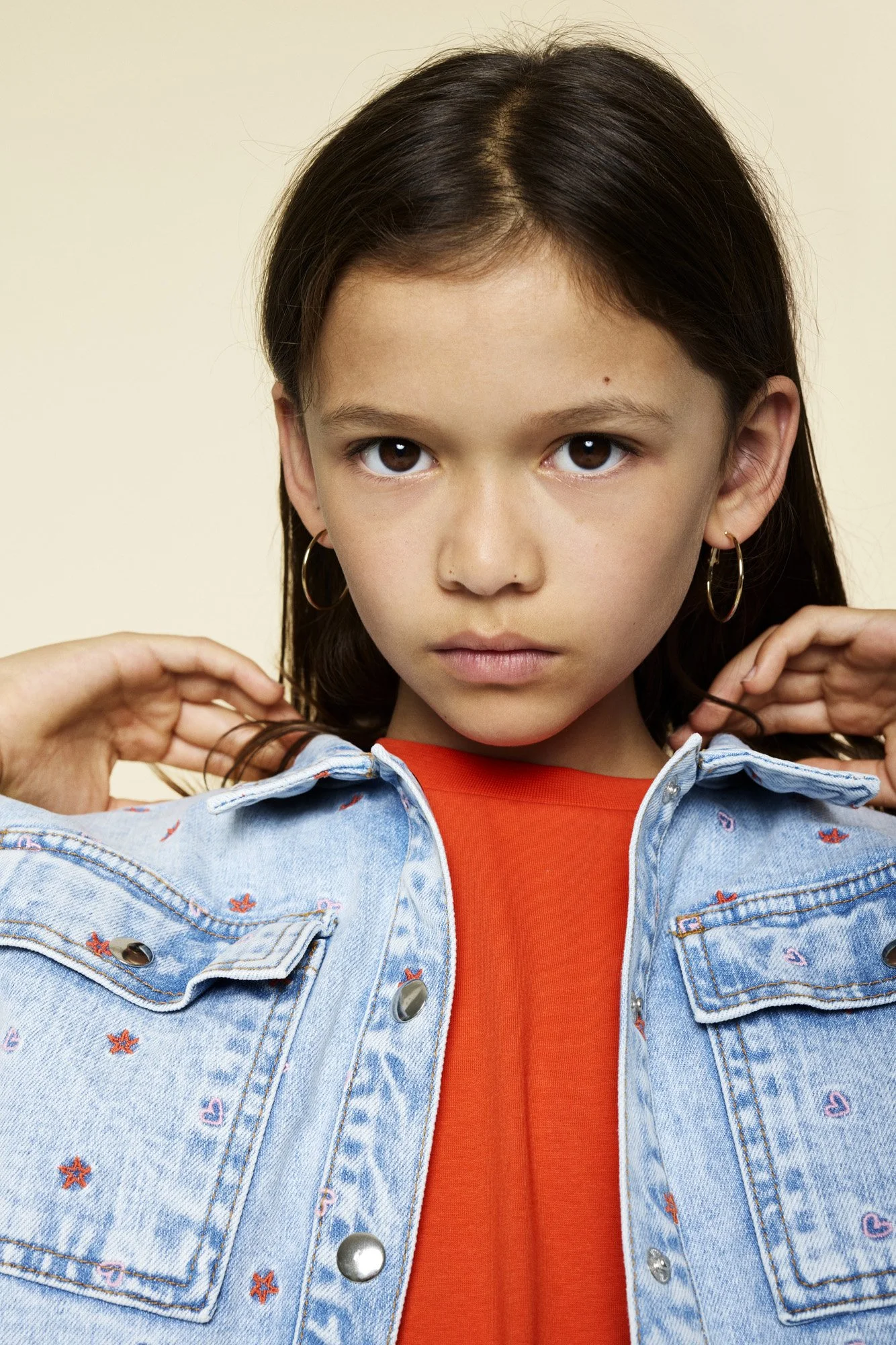 A young girl with brown hair, hoop earrings, and a serious expression, wearing a denim jacket with embroidered designs over an orange shirt.