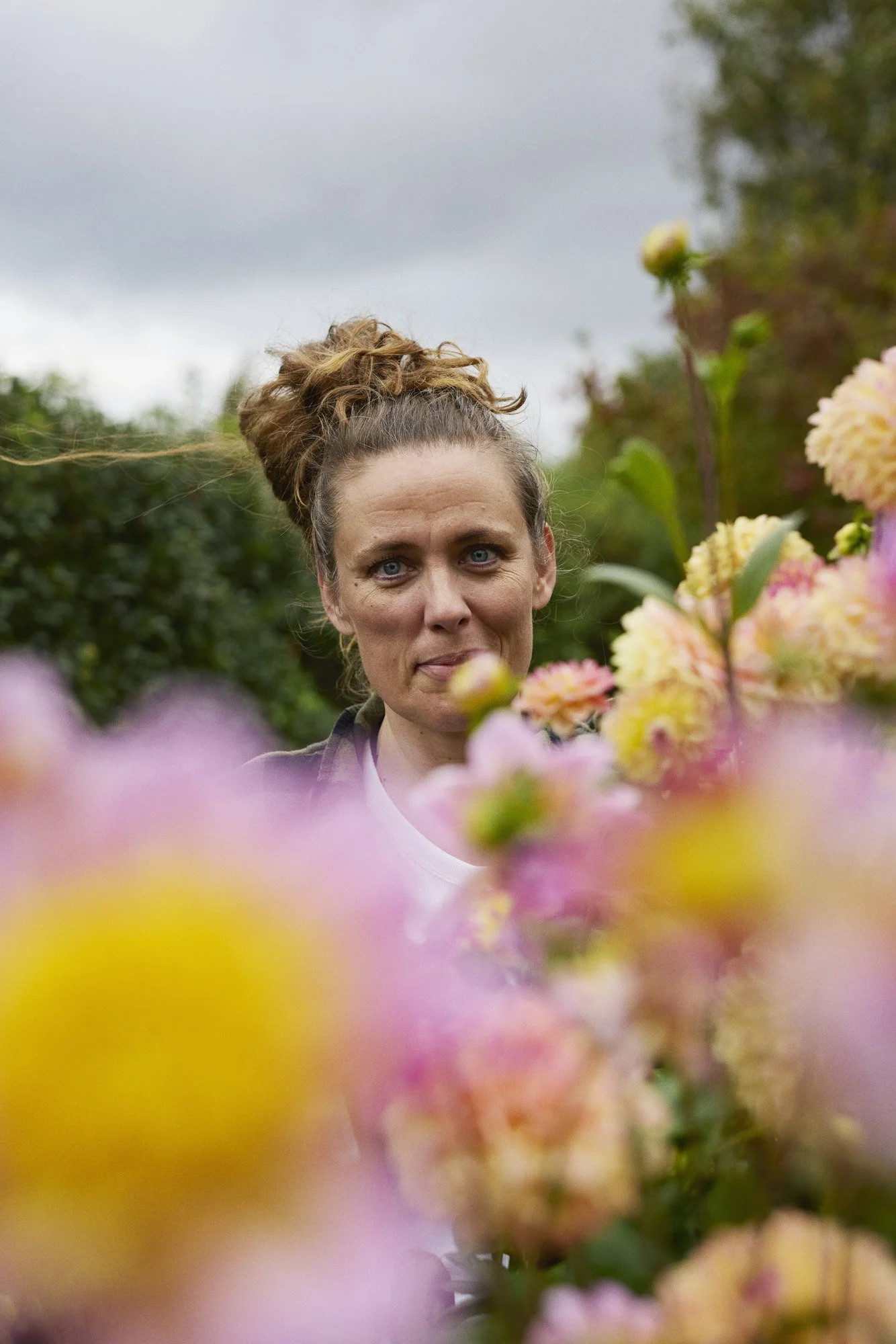 Cristine Glise - Haven Håbets og Kærligheden A woman with a curly bun hairstyle and light skin looks at the camera among pink and yellow flowers outdoors on a cloudy day.