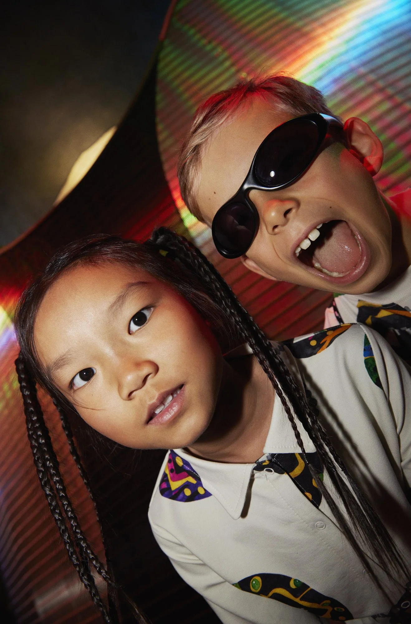 Two children, one with braided hair and the other wearing sunglasses, posing with a large, colorful, iridescent umbrella against a dark background.