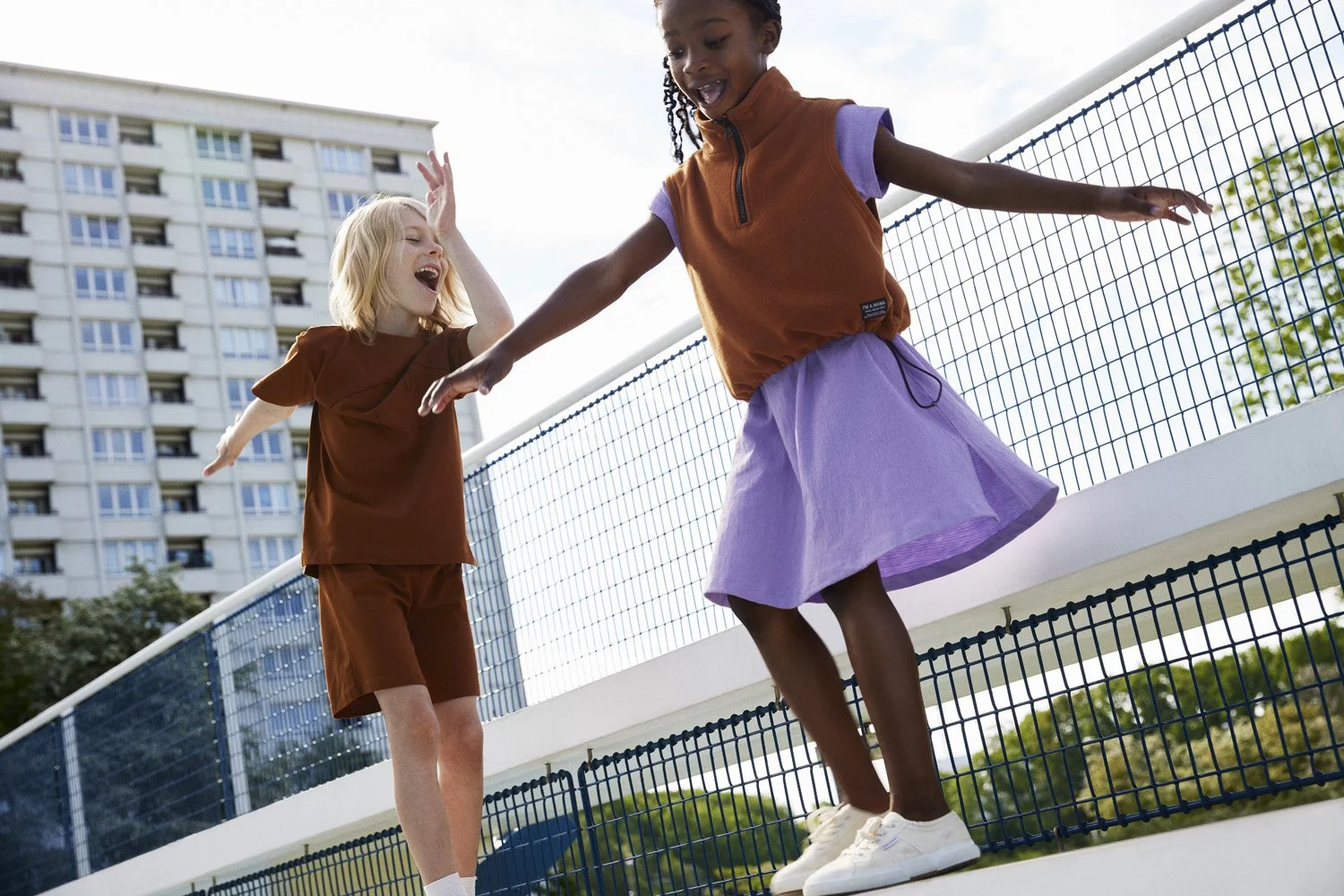 Two young girls playing on a skate park with a high-rise building in the background. The girl on the left has blonde hair, is laughing, and has her hand raised. The girl on the right has braided hair and is balancing on the edge of the skate ramp.
