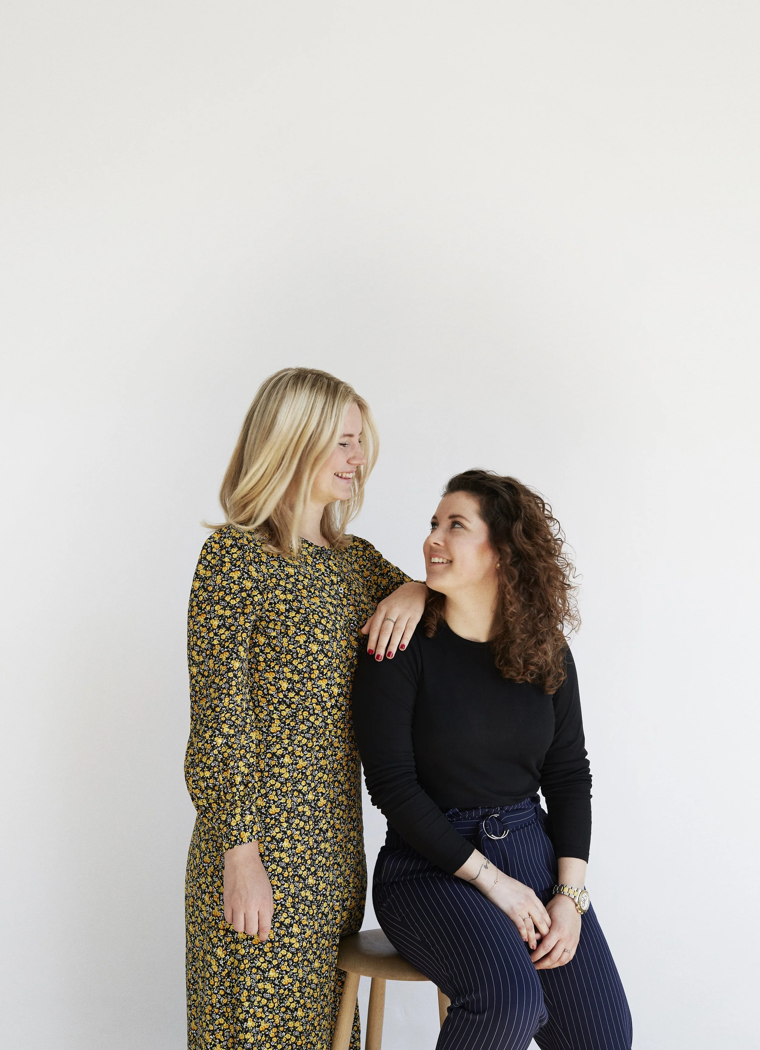 Two women smiling at each other against a plain white wall. One woman is seated on a stool, wearing a black top and striped pants, while the other stands beside her, wearing a yellow floral dress.