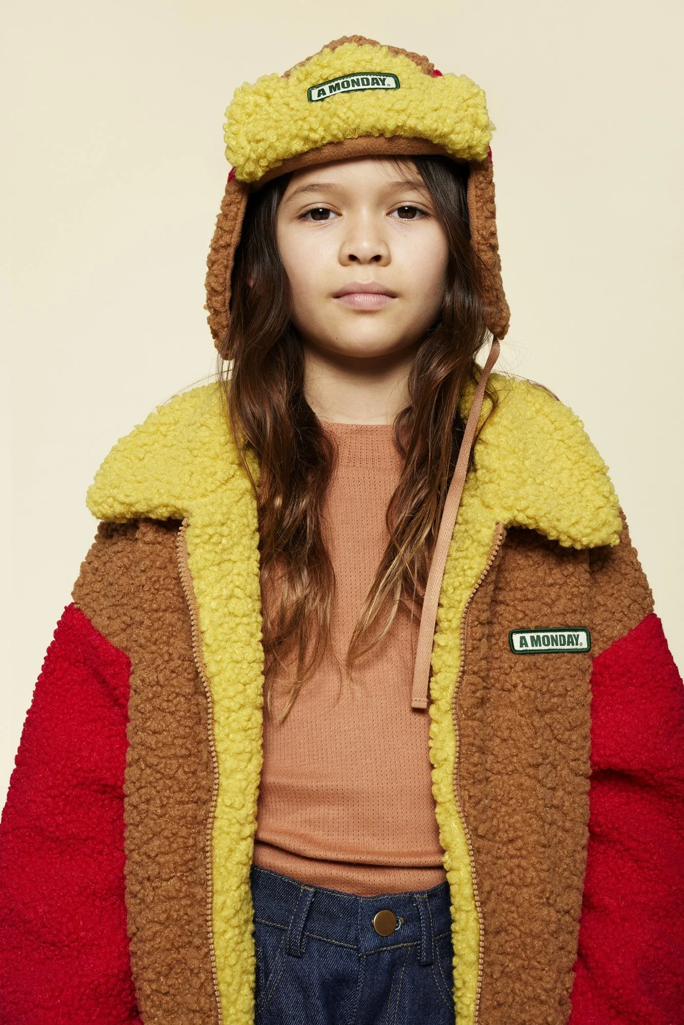 A young girl wearing a colorful, textured fleece jacket and a cozy trapper hat with the words "A MONDAY" on the front, standing against a plain light-colored background.