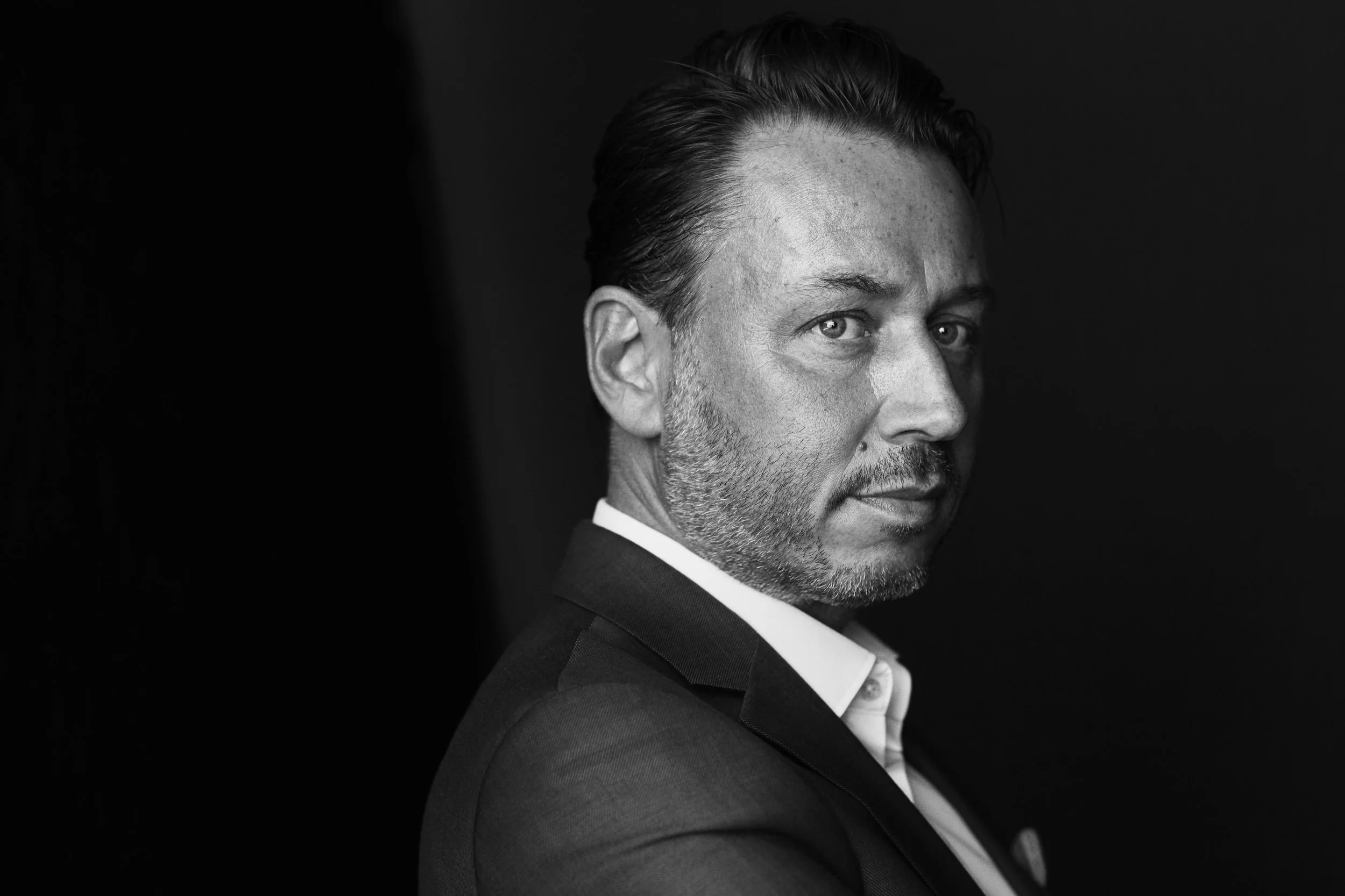 Black and white portrait of a middle-aged man with short, slicked-back hair, a beard, and piercing eyes, wearing a suit and white shirt, looking over his shoulder against a dark background.