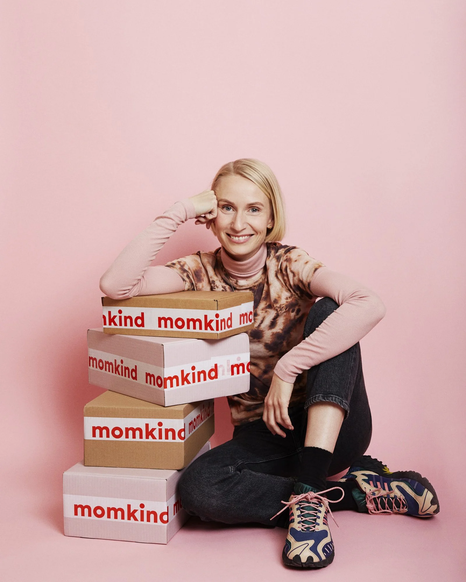 Portrait Nanna Schultz founder Momkind - A smiling woman with short blonde hair sitting on the floor next to a stack of four boxes with "momkind" written on them against a pink background. She is wearing a tie-dye shirt, pink long-sleeve shirt undern