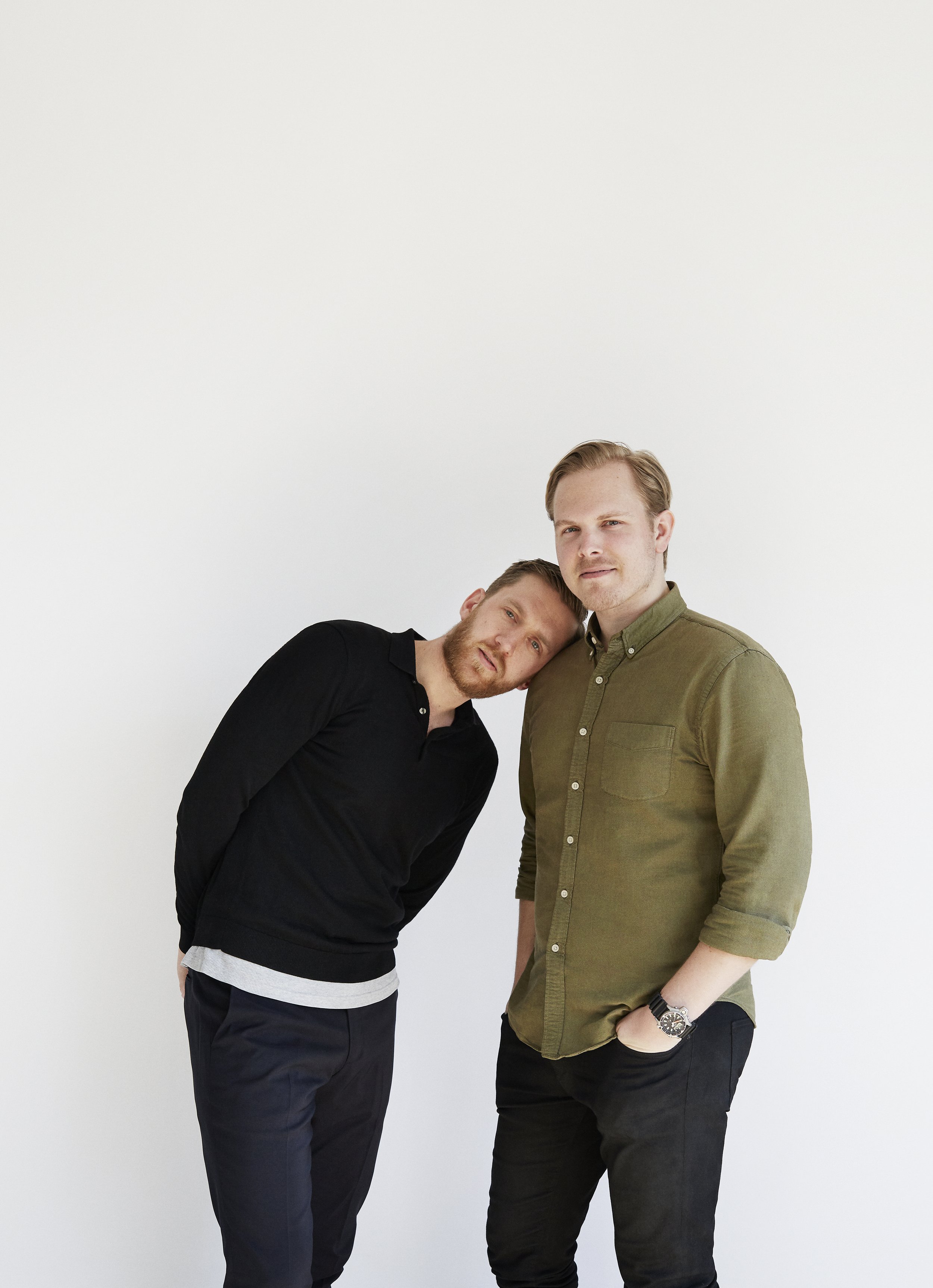 Two young men standing close together against a plain white background. One man leans his head on the other's shoulder. Both are casually dressed, with the man on the left in a black sweater and the man on the right in an olive green shirt.