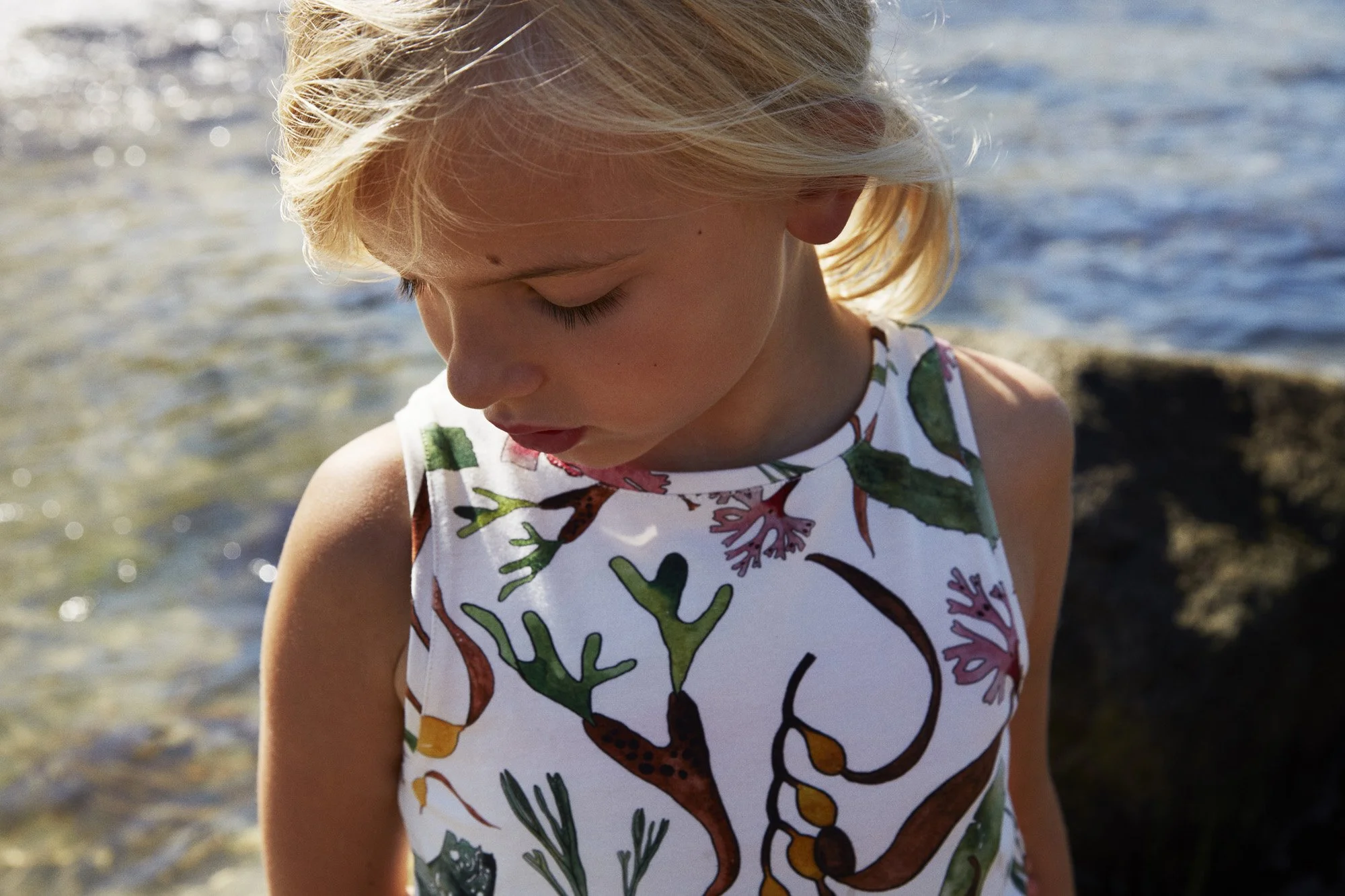 A young girl with blonde hair looking down at the water near a rocky shoreline, wearing a sleeveless dress with a botanical pattern.