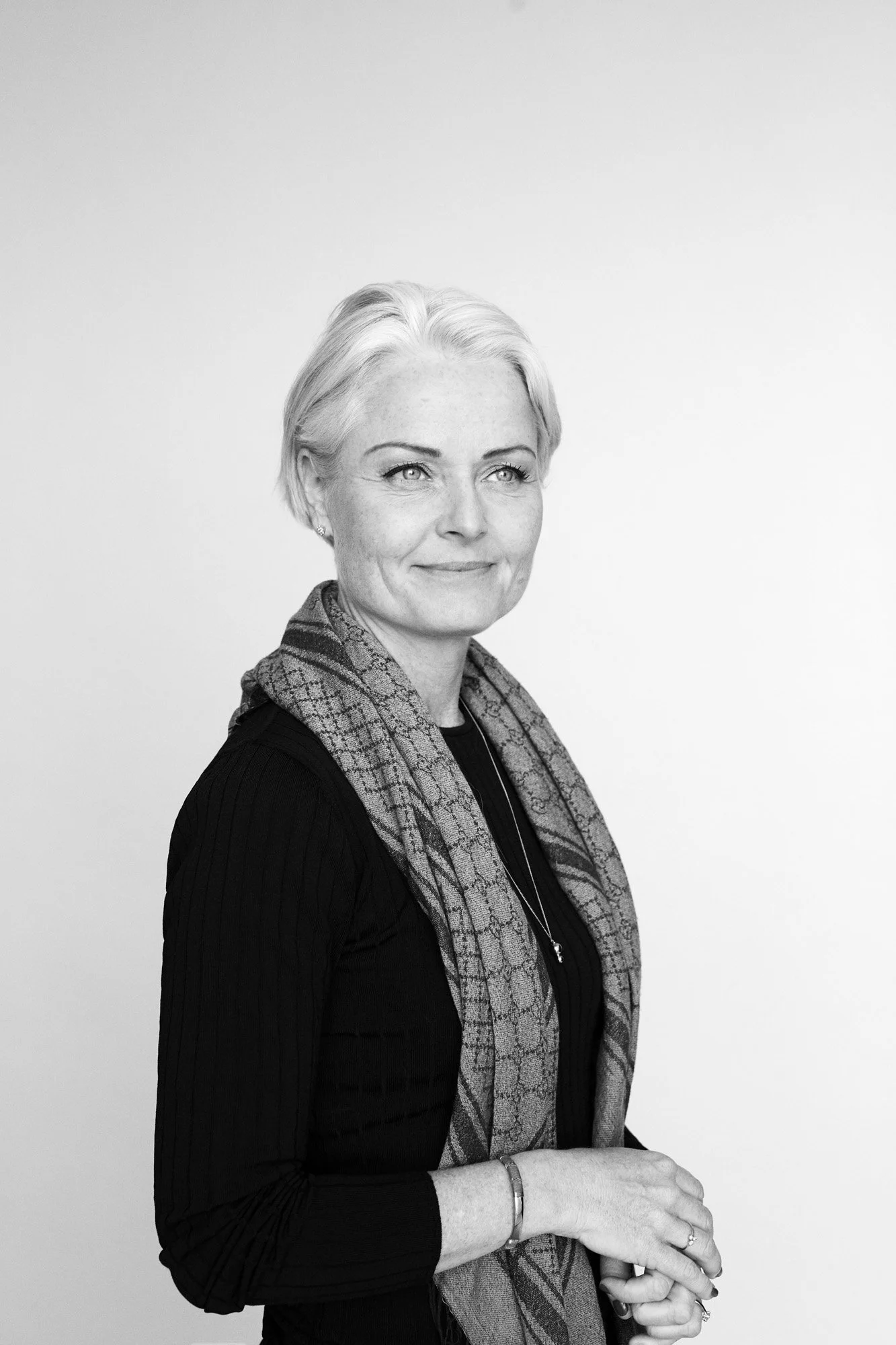 Black and white portrait of a woman with short, light-colored hair, wearing a dark top and a patterned scarf, standing against a plain background.