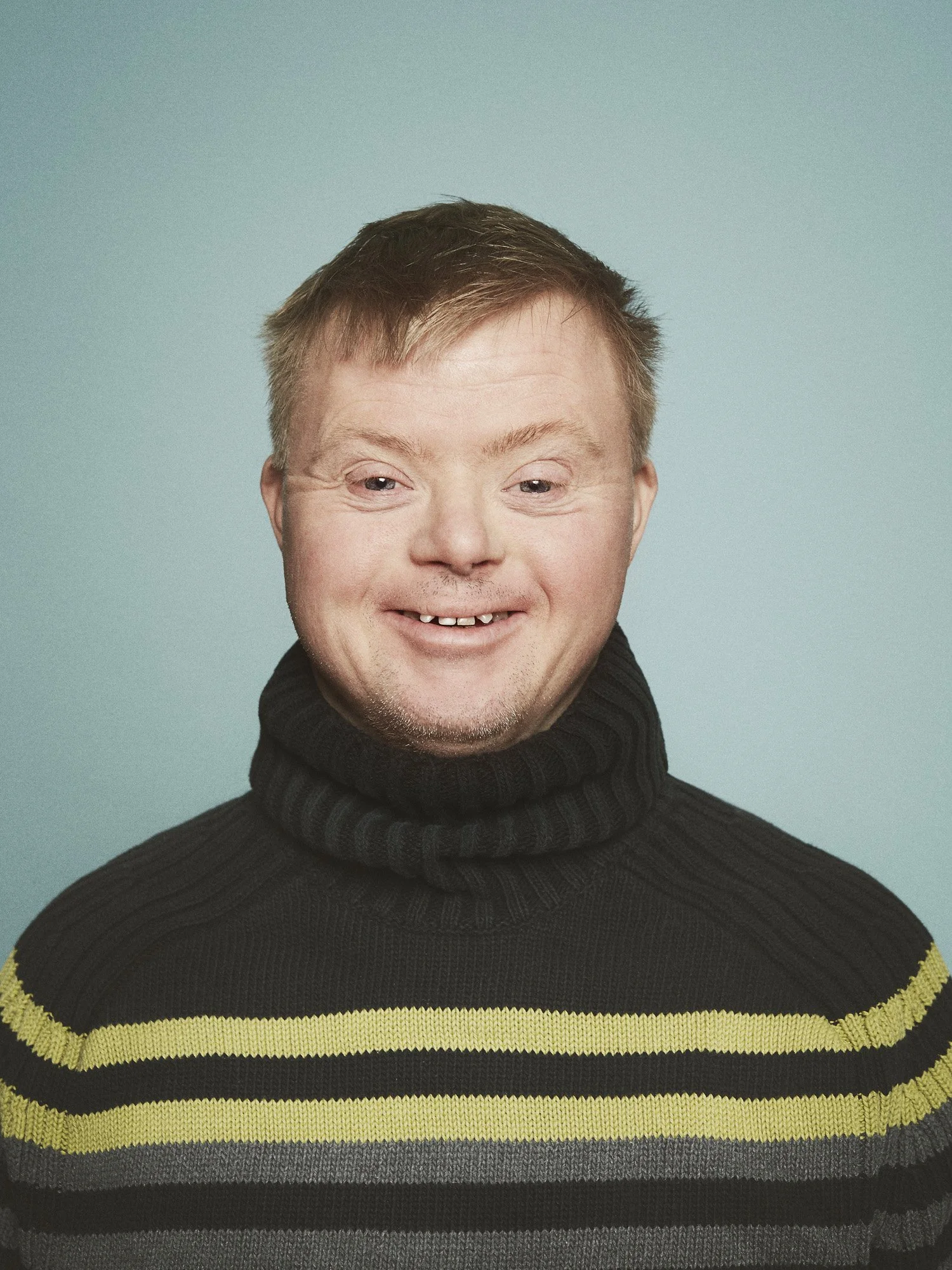 GLAD Teater - A smiling man with short light brown hair, wearing a black turtleneck sweater with yellow and gray stripes, posing against a plain teal background.