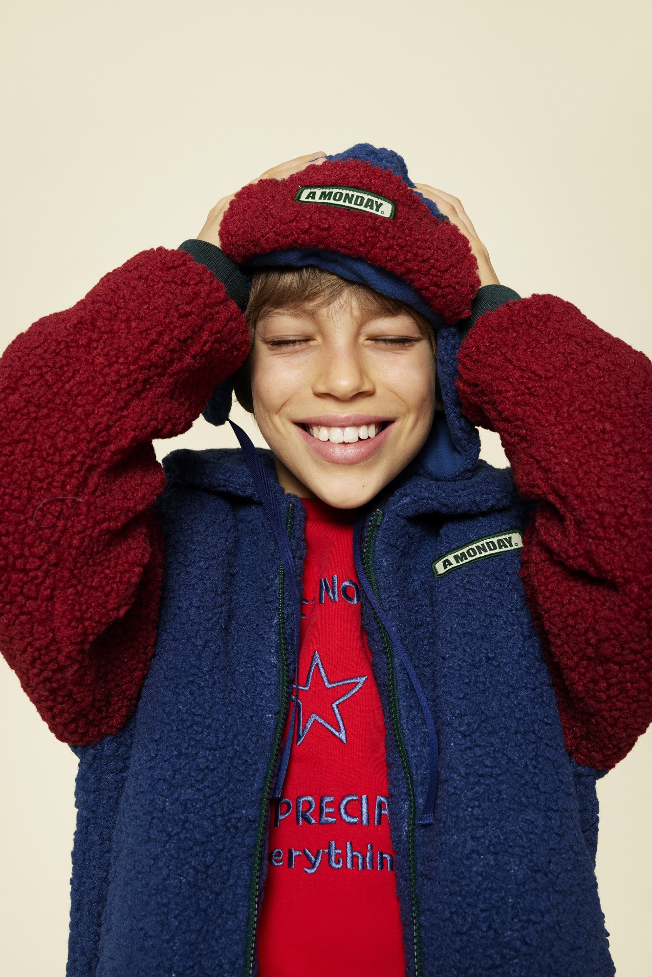 Smiling boy wearing a red and blue beanie and a blue and red fleece jacket, holding the beanie with both hands.