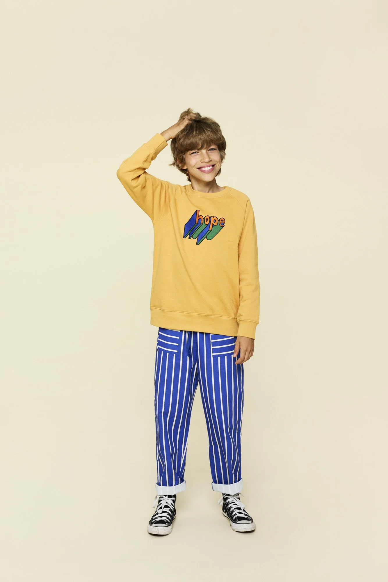 A young boy wearing a yellow sweater with 'hope' written on it, blue striped pants, and black high-top sneakers, standing against a plain background, smiling, with one hand on his head.