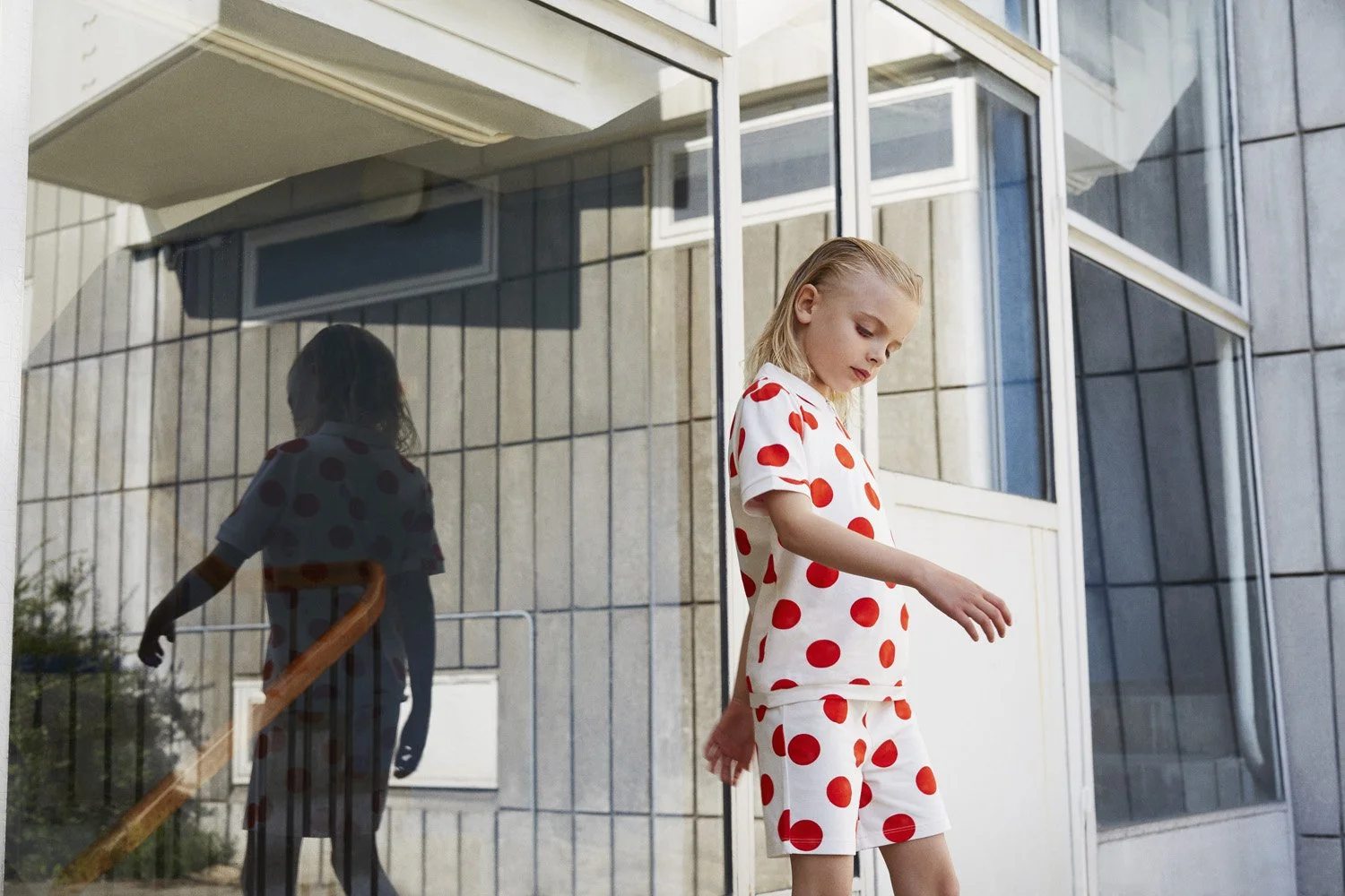 WAWA CPH - Young girl in white pajamas with red polka dots standing outside near a glass building, with her reflection visible in the glass.