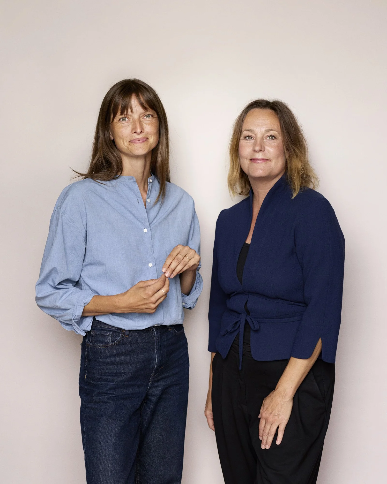 Two women standing side by side against a plain light-colored background, looking at the camera. One woman has shoulder-length brown hair, wearing a light blue button-up shirt and dark jeans. The other woman has shoulder-length blonde hair, wearing a