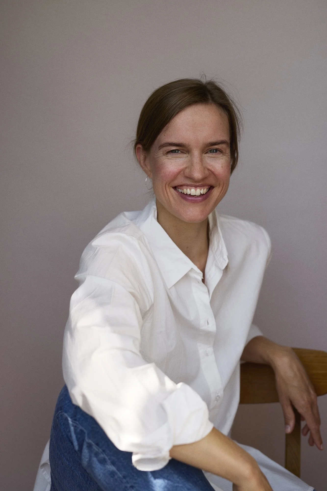 A woman with brown hair, wearing a white button-up shirt, smiling and sitting on a wooden chair with a plain gray background.