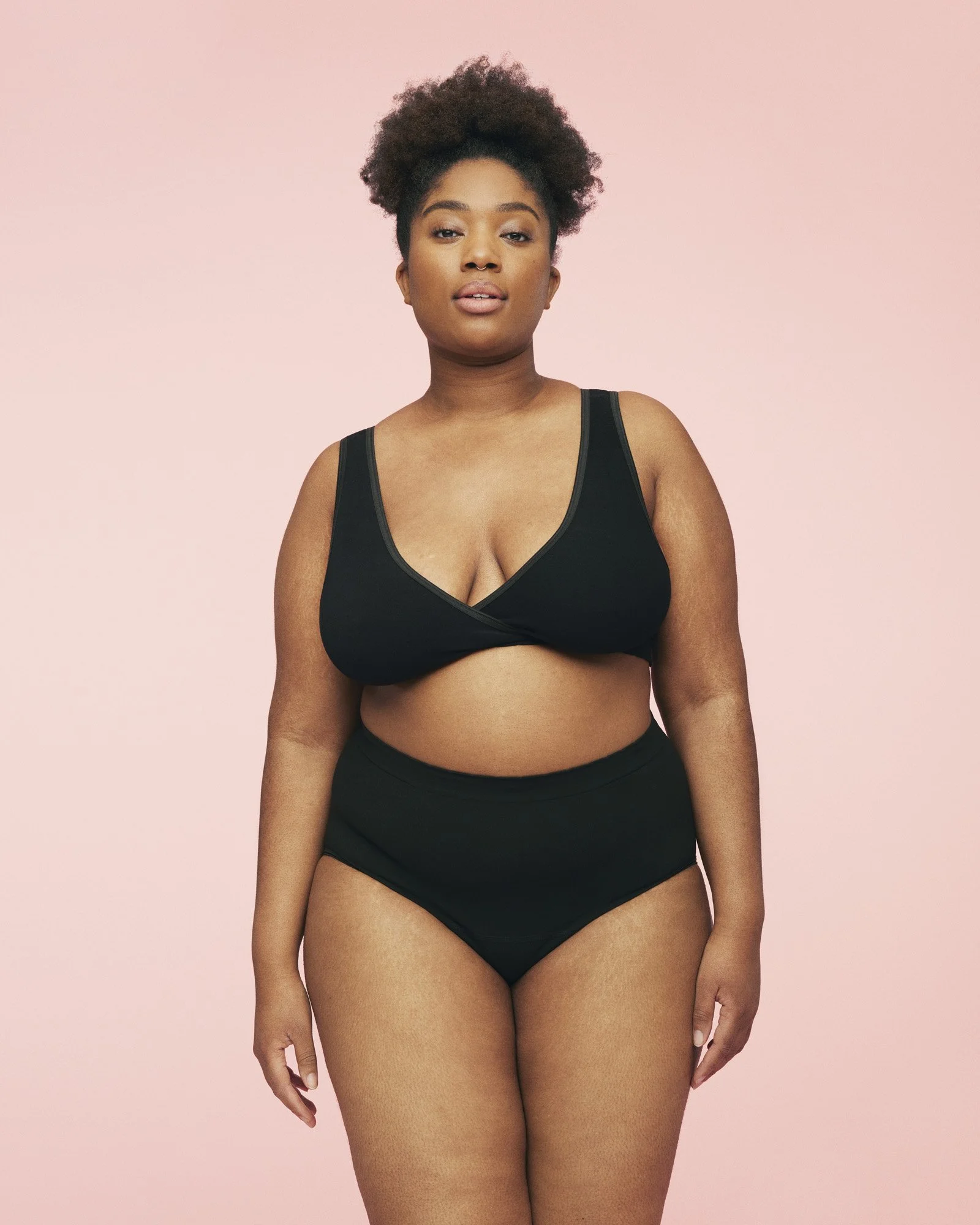 MOMKIND - A woman with short curly hair wearing a black bralette and high-waisted black underwear standing against a pink background.