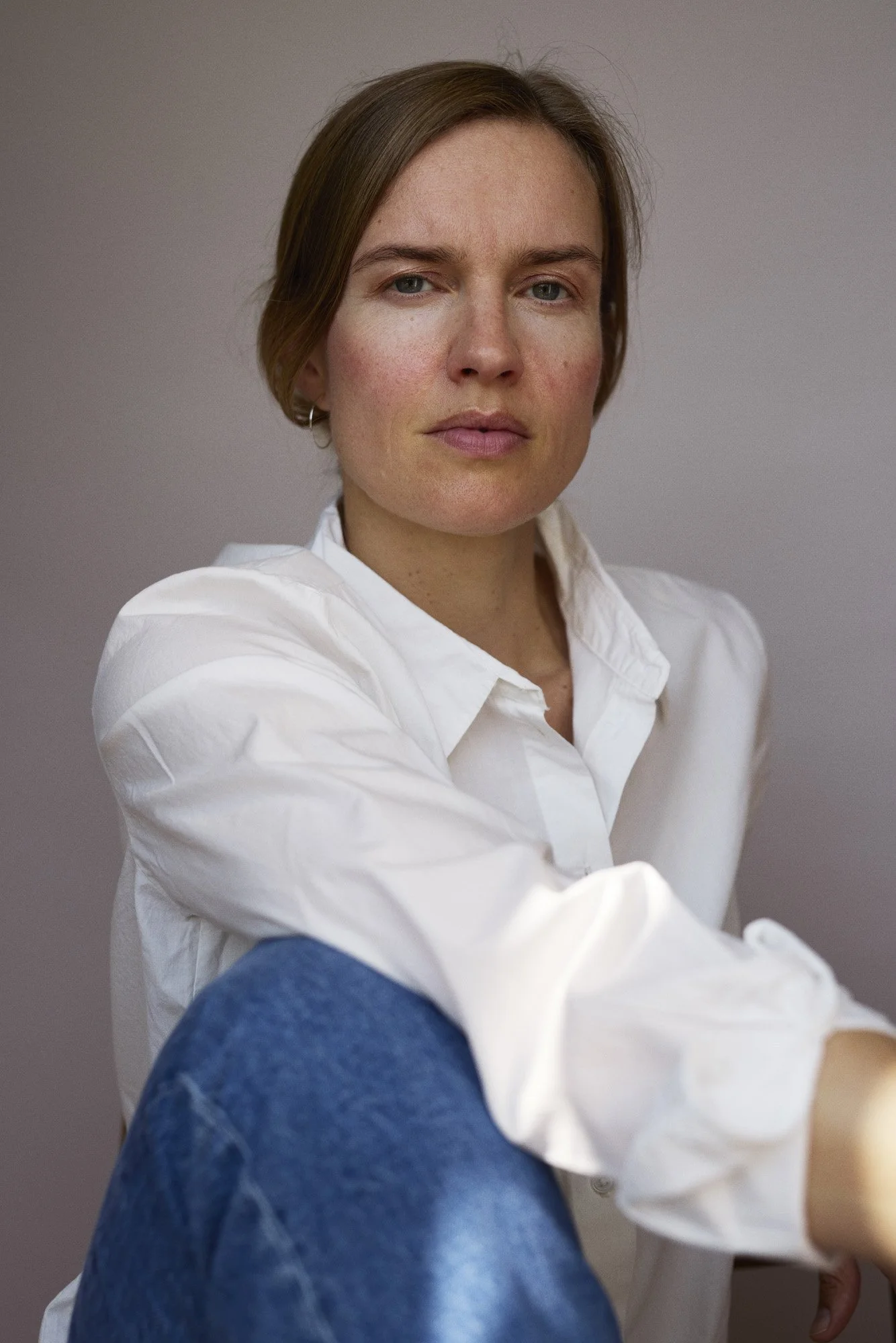 A woman with shoulder-length brown hair, wearing a white shirt and blue jeans, sitting against a plain gray background.