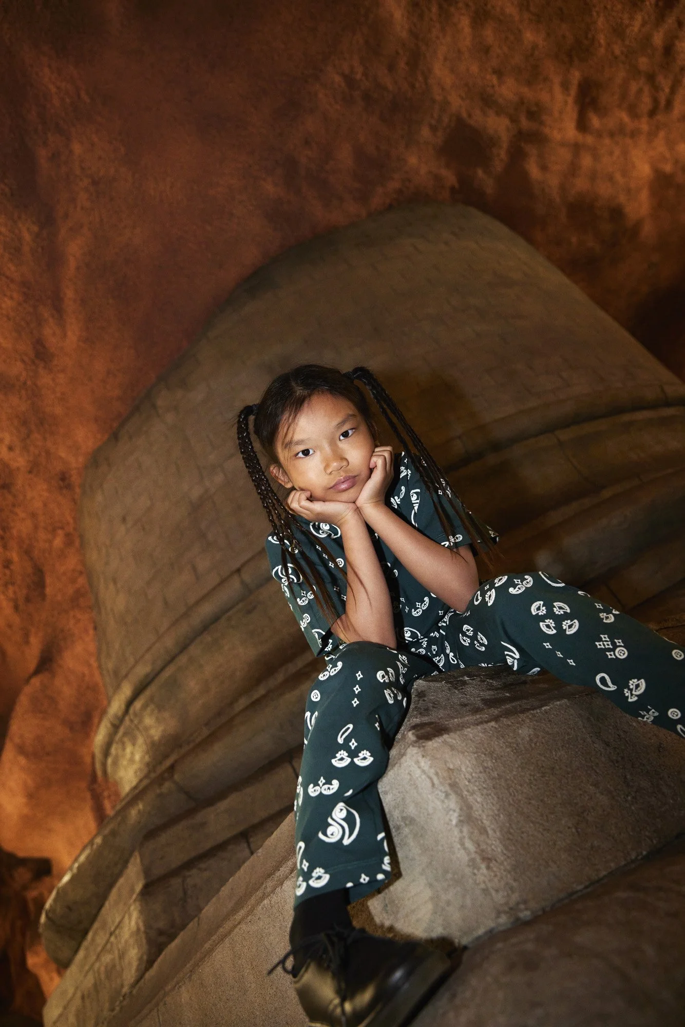 Young girl with braided hair sitting on a stone ledge in a cave, resting her chin on her hands, wearing a matching dark green patterned outfit.