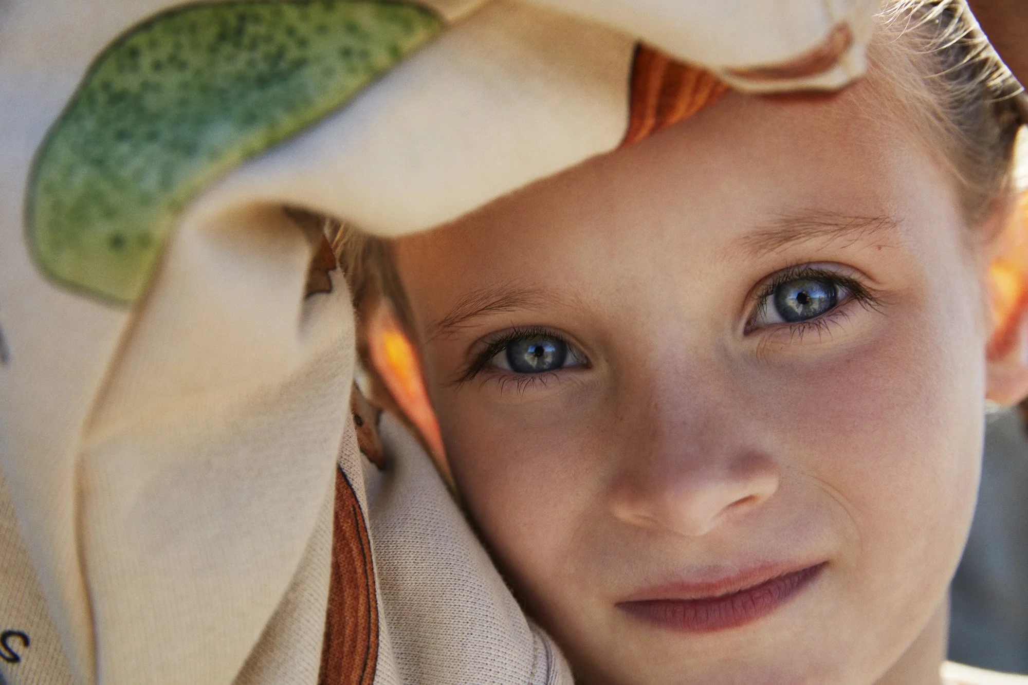 Close-up of a young boy with blue eyes and blonde hair, resting his head on a person's shoulder, with a patterned fabric partially covering his head.