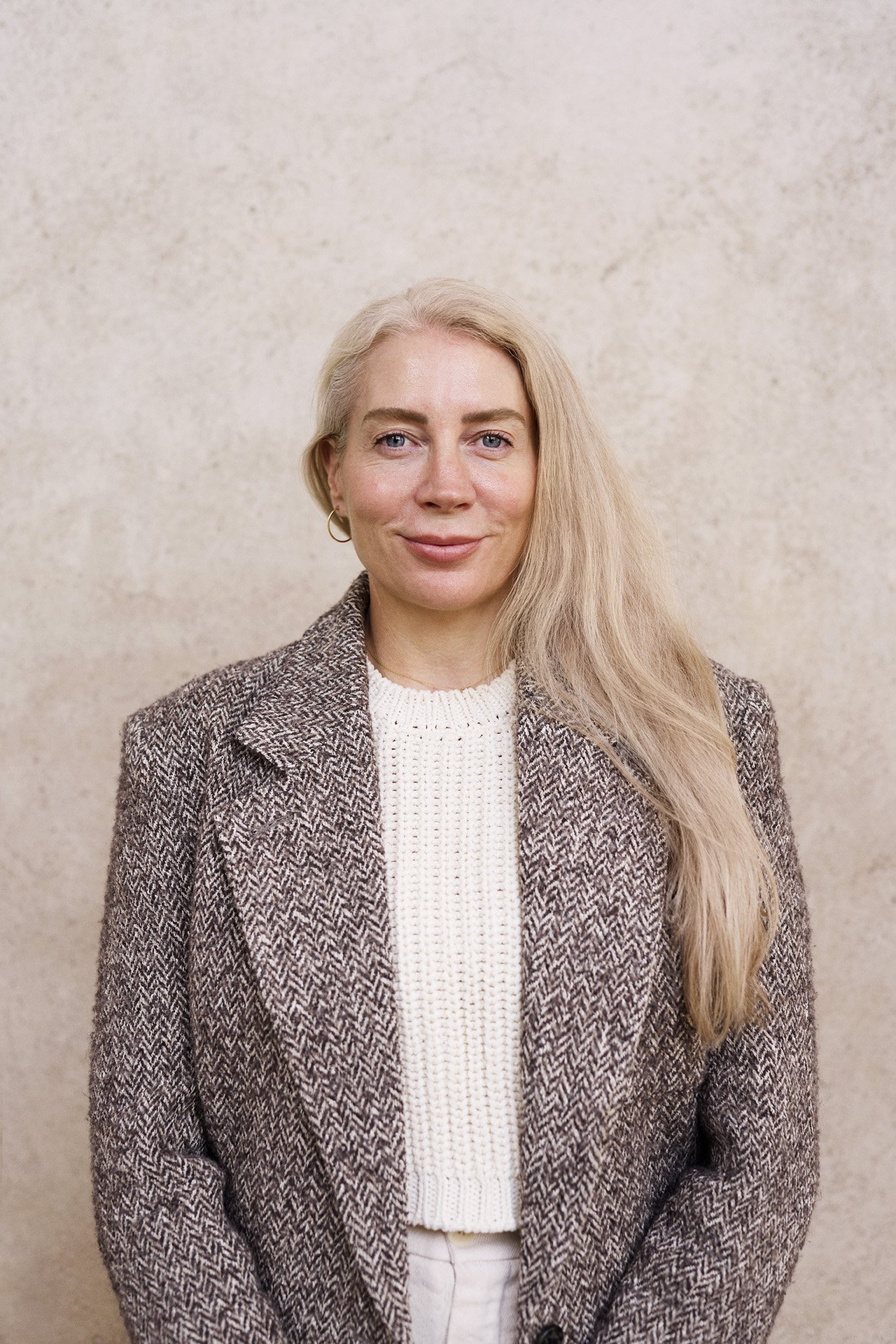 Portrait  Unconventional Ventures - Girl with long blonde hair wearing a brown patterned blazer and a white knitted sweater, standing against a beige textured background.