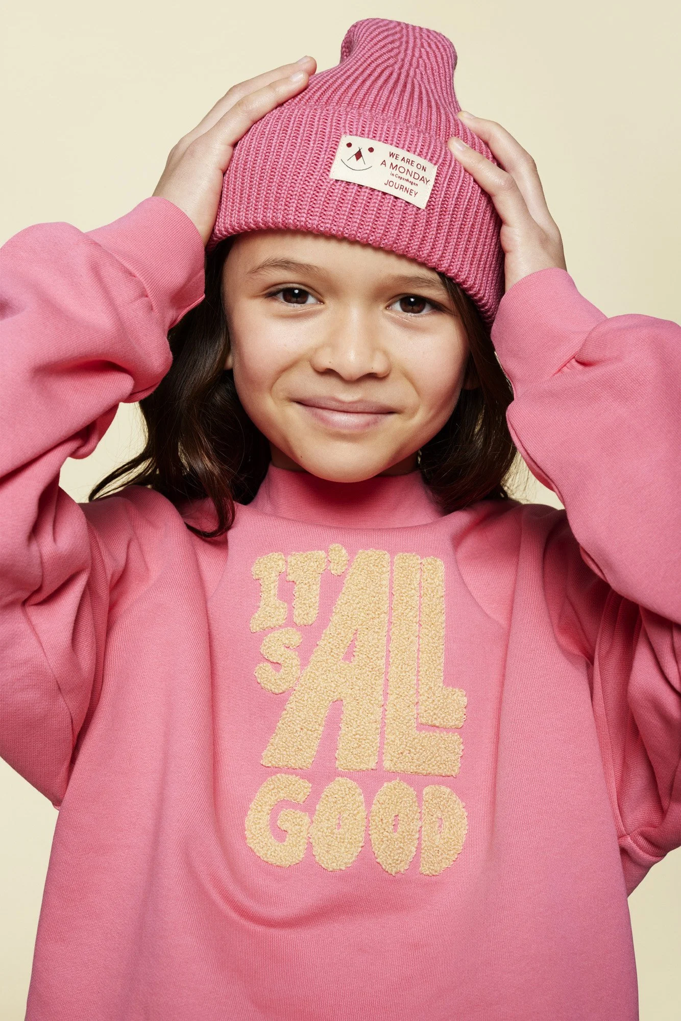 A young girl with brown hair wearing a pink beanie and pink sweatshirt with 'IT'S ALL GOOD' written on it, smiling at the camera.