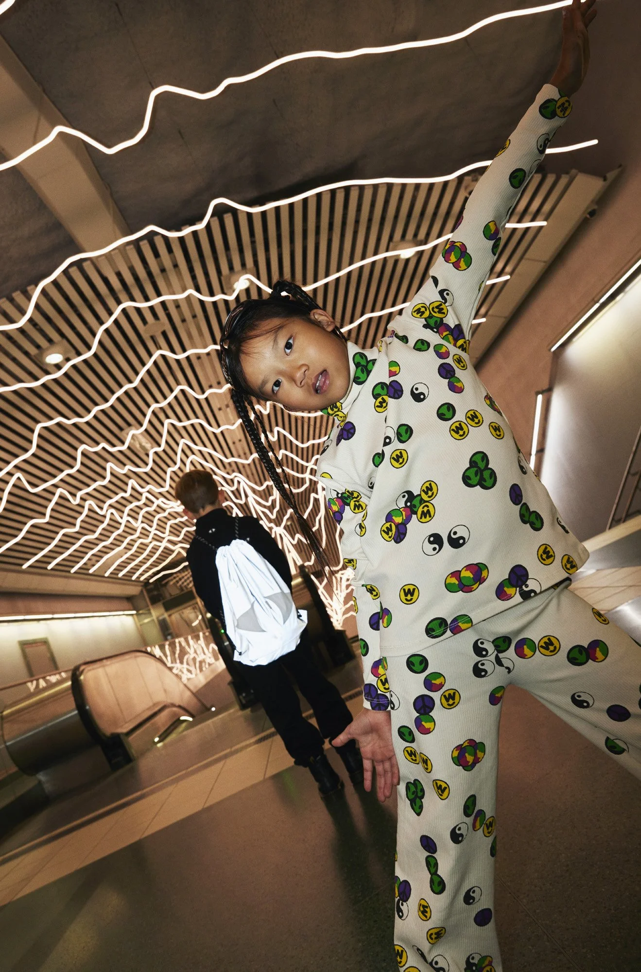 A girl in matching yin-yang and colorful circle patterned pajamas stretching with one arm raised at an indoor location with artistic light fixtures on the ceiling.