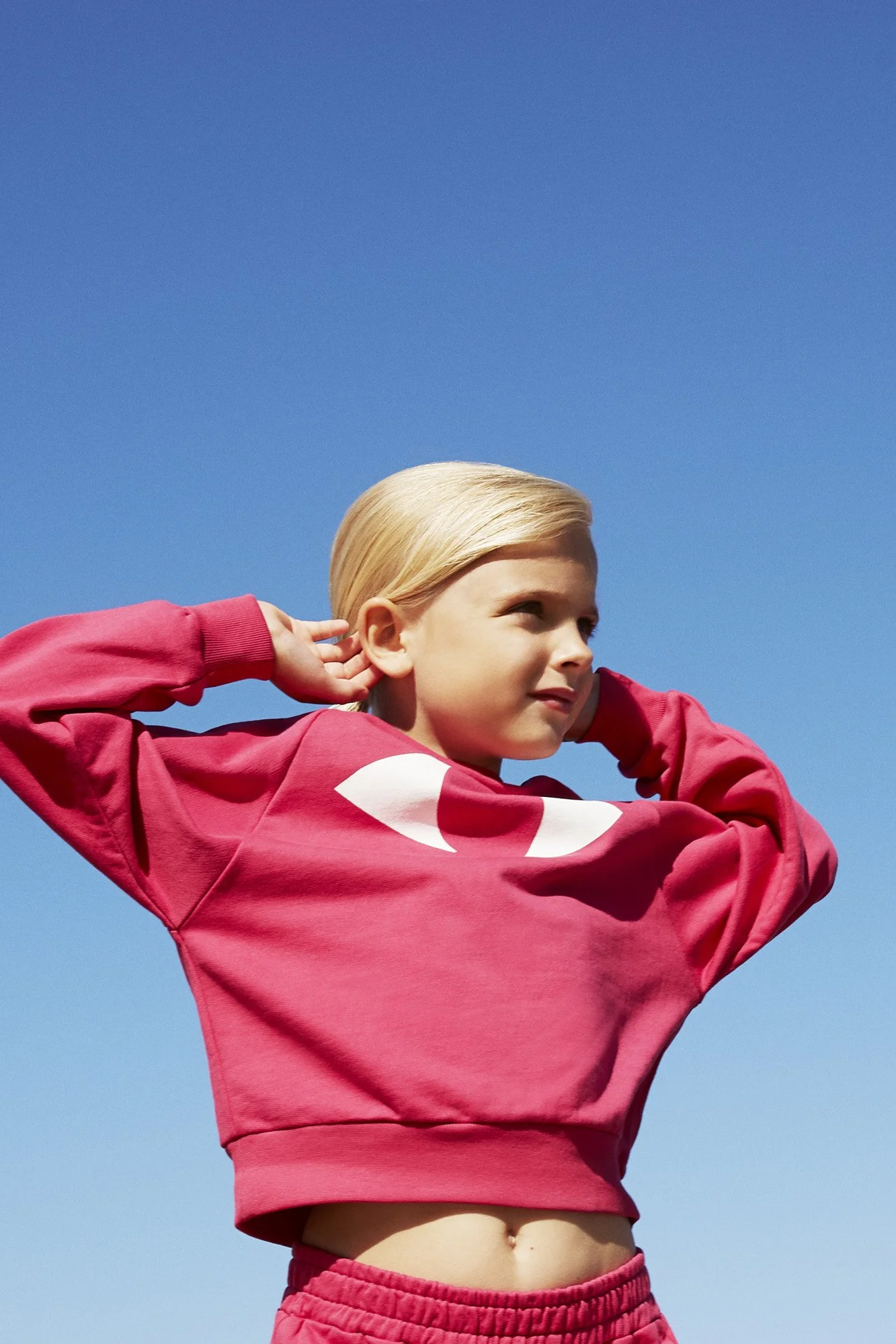 A young girl with blonde hair wearing a pink sweatshirt and matching pink shorts, standing outdoors against a bright blue sky. She is touching her ears and looking off to the side.