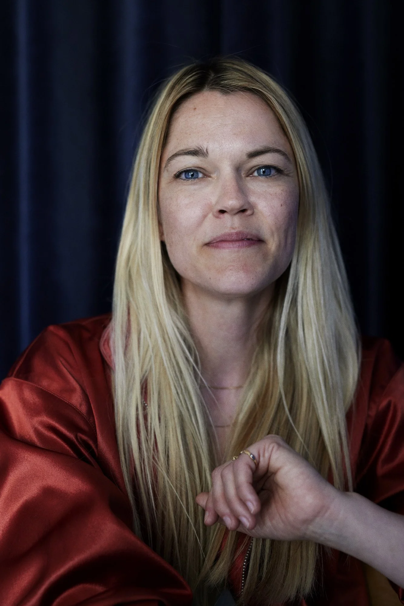 Portrait Psykoterapeut Lena Breum - Portrait of a woman with long blonde hair, blue eyes, and light skin, wearing a red satin jacket, sitting in front of a dark background.