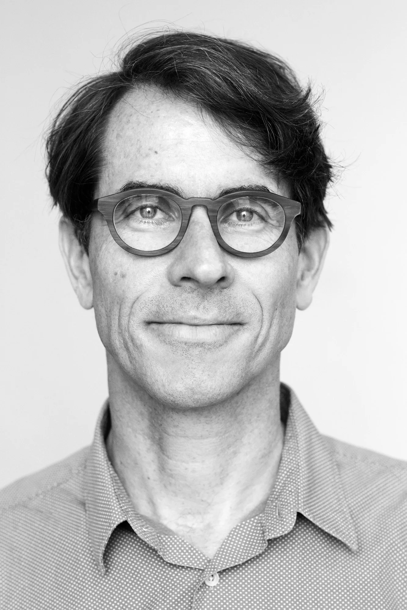 Black and white portrait of a man wearing glasses and a patterned shirt, smiling softly.
