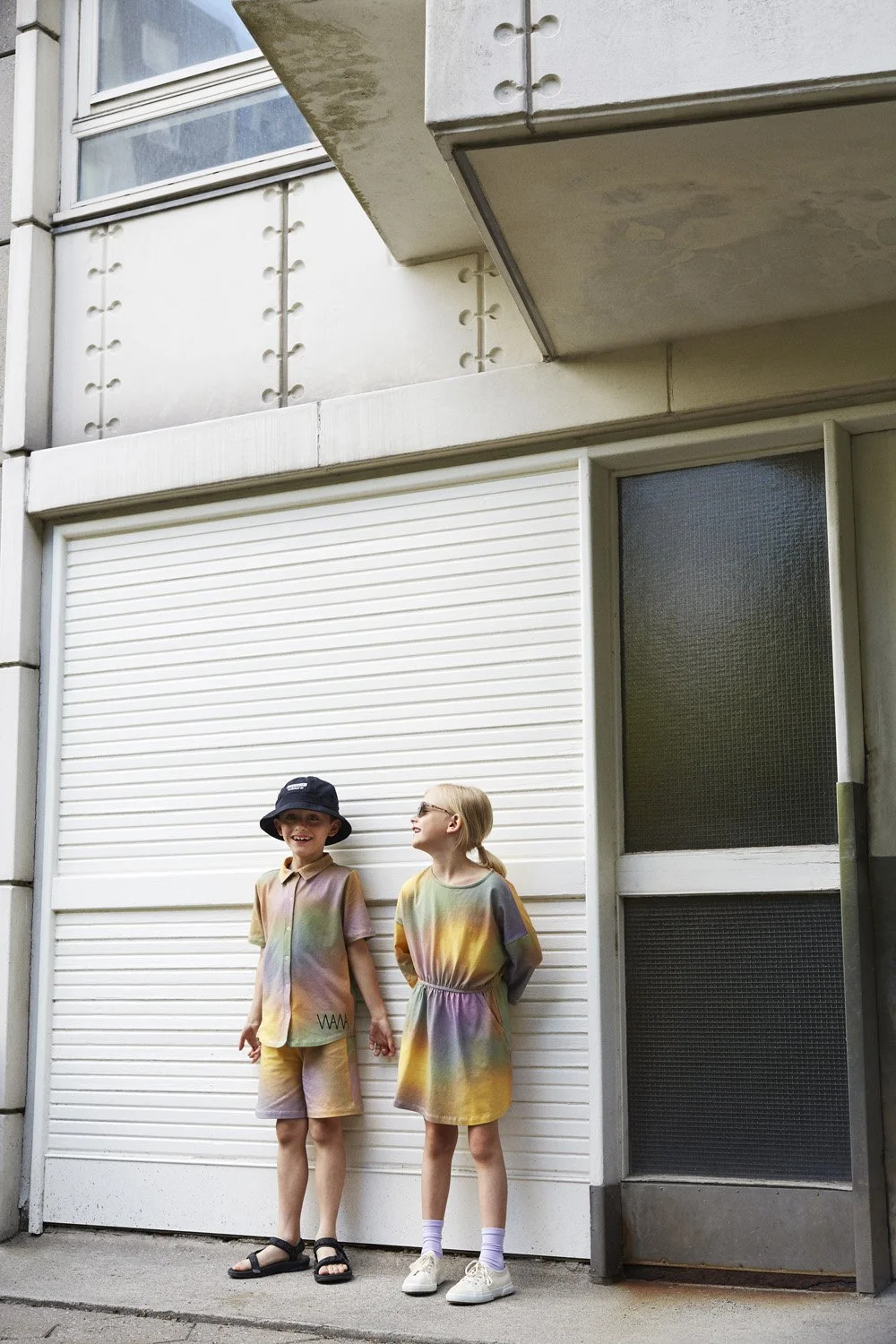 WAWA CPH - Two children standing outside a building in front of a white roll-up door, holding hands, wearing matching rainbow-colored outfits, smiling and looking at each other.