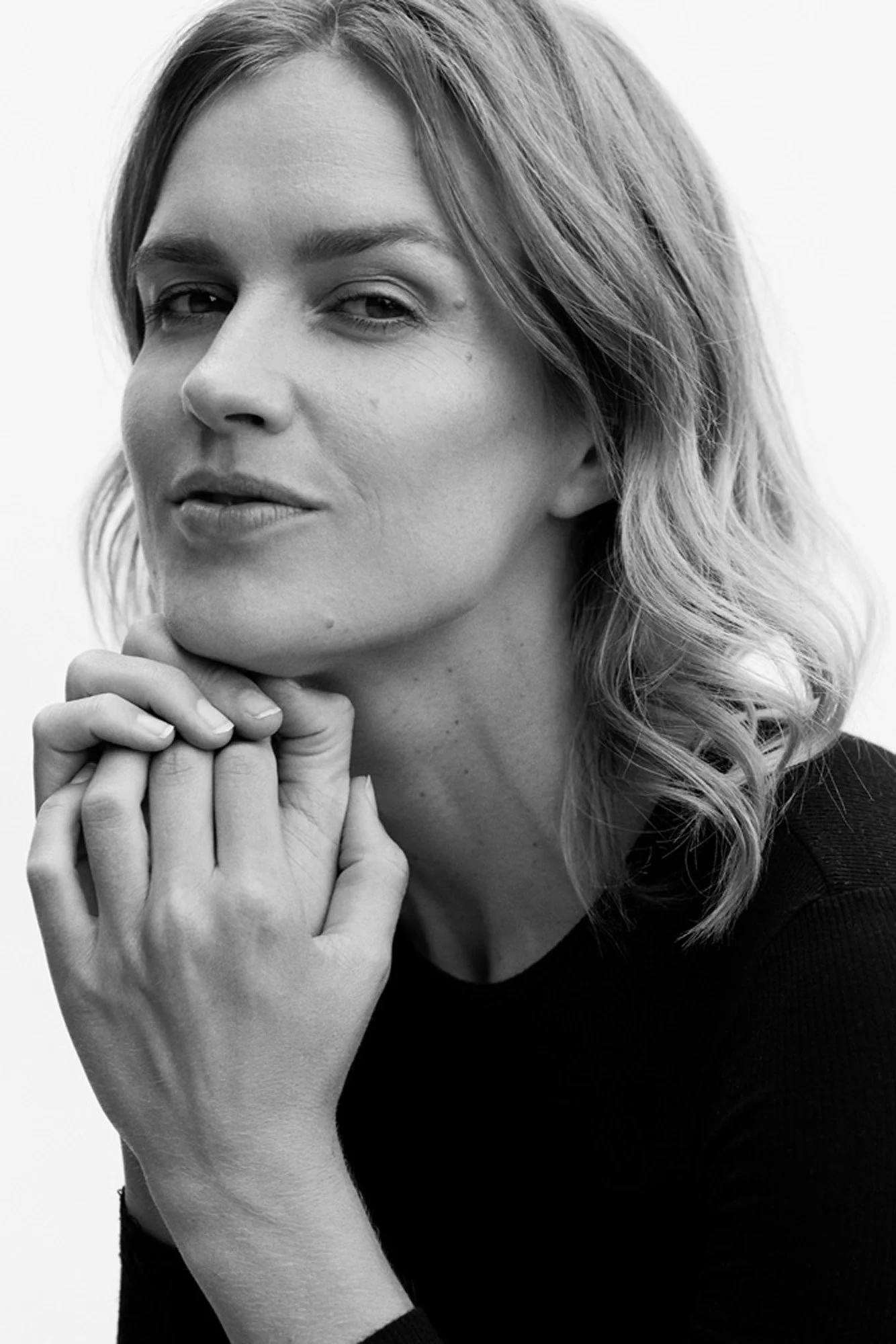 Black and white portrait of a woman with wavy hair, resting her chin on her hands, looking at the camera.