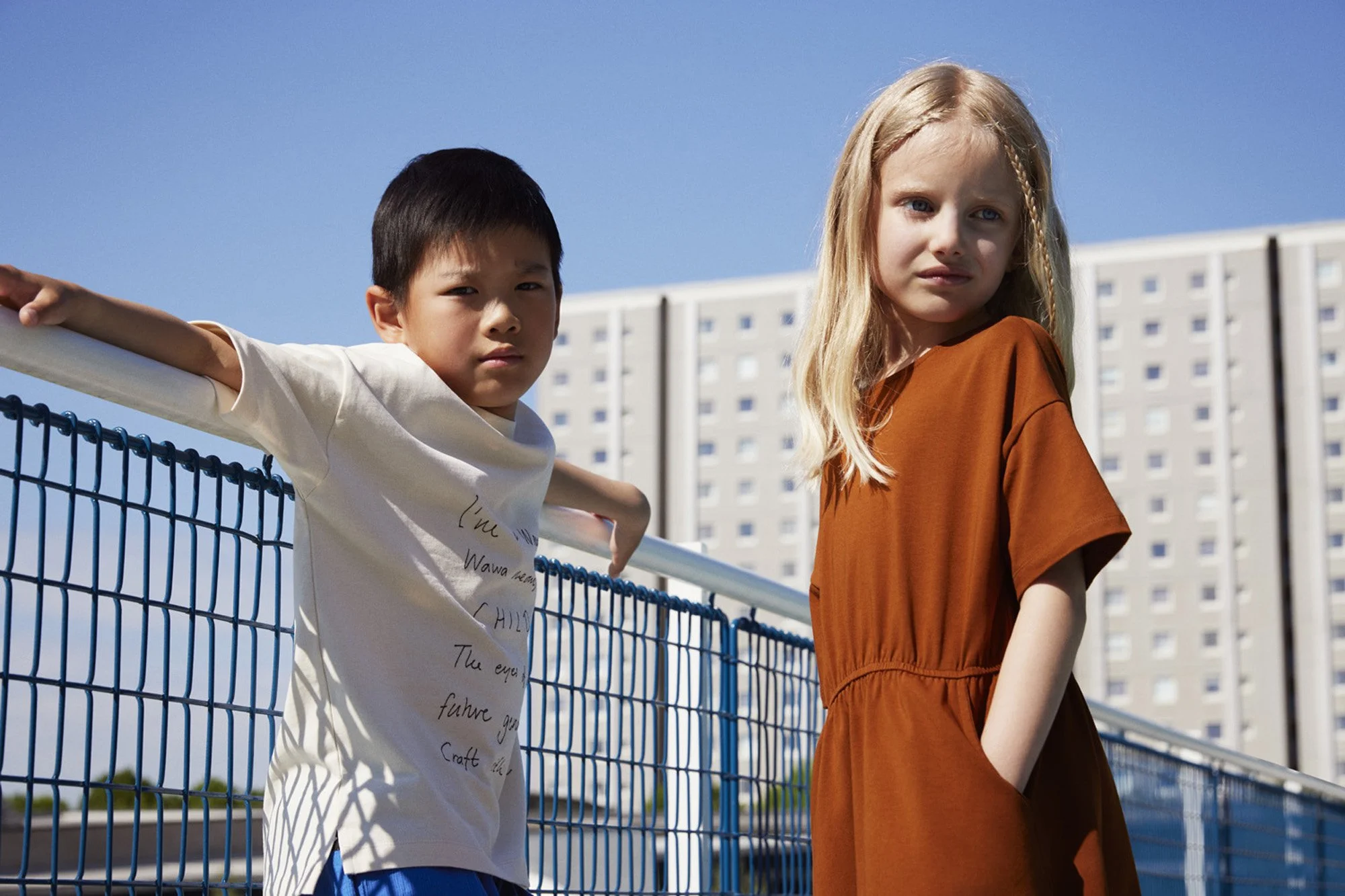 WAWA CPH - Young boy and girl standing near a blue tennis court fence with a high-rise building in the background on a sunny day.