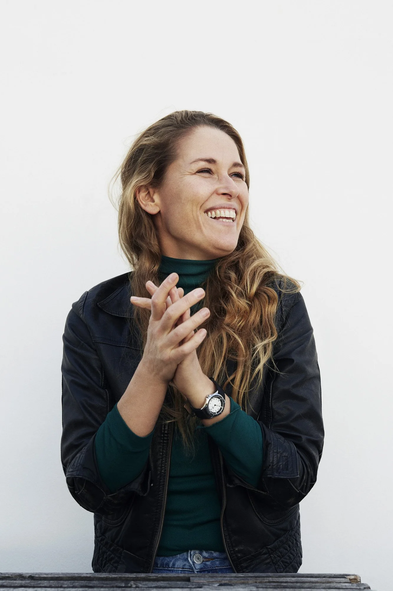 Portrait Lea Korsgaard Zetland - A woman with long, wavy, light brown hair smiling and clapping her hands, wearing a black leather jacket, green turtleneck, watch, and jeans, against a plain white background.