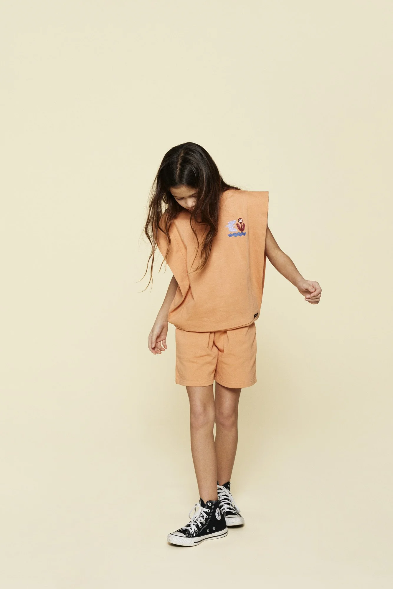 A young girl with long brown hair wearing a peach-colored sleeveless top and shorts with black high-top sneakers, standing against a plain light background.