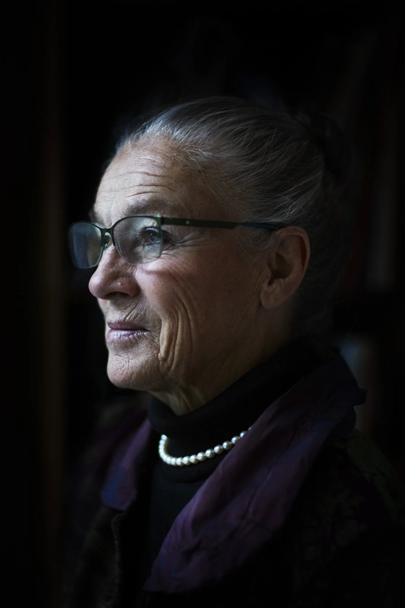 Portrait Ritt Bjerregaard - Side profile of an elderly woman with glasses, gray hair in a bun, wearing a pearl necklace and dark clothing, against a dark background.