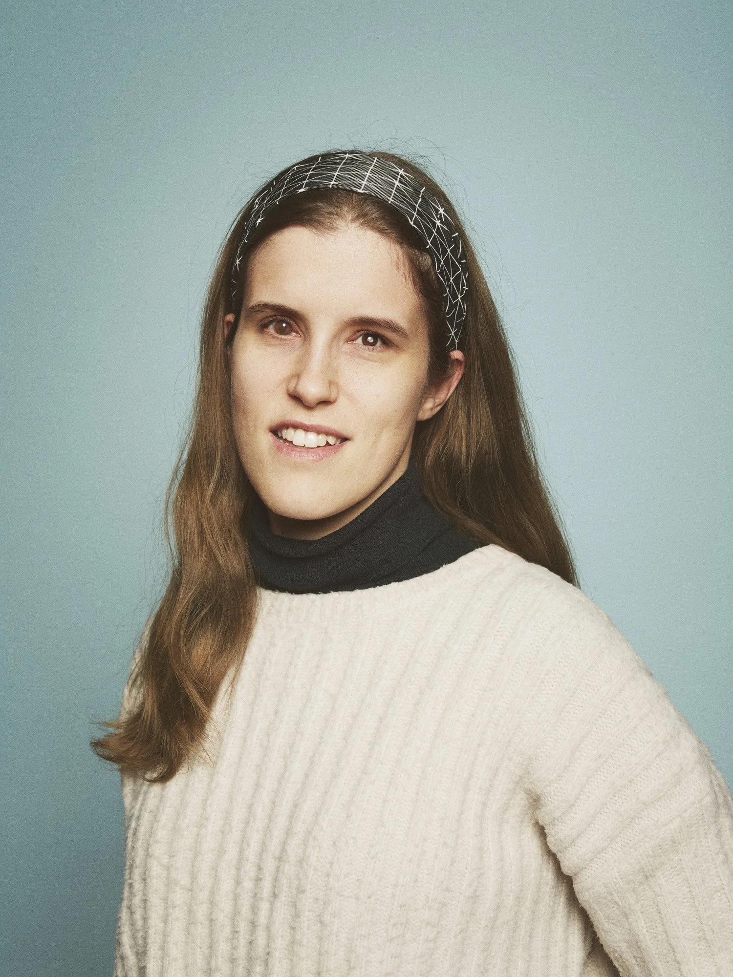 GLAD Teater - Portrait of a young woman with long brown hair, wearing a black turtleneck under a cream sweater, and a patterned headband, against a blue background.