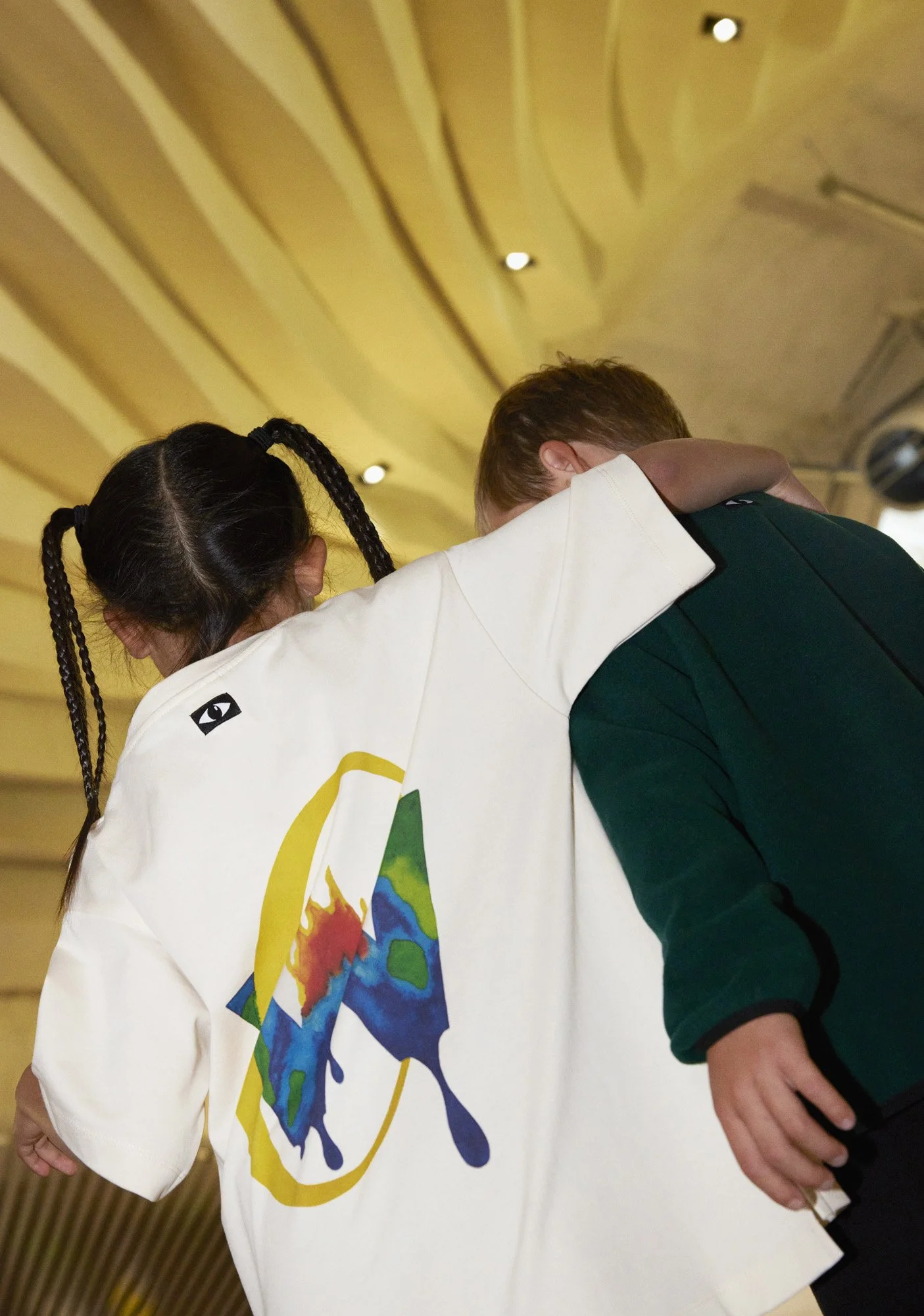 A girl with braided hair wearing a white coat with a colorful abstract design, hugging a boy with short hair dressed in a dark green jacket, inside a room with a curved wooden ceiling and small ceiling lights.