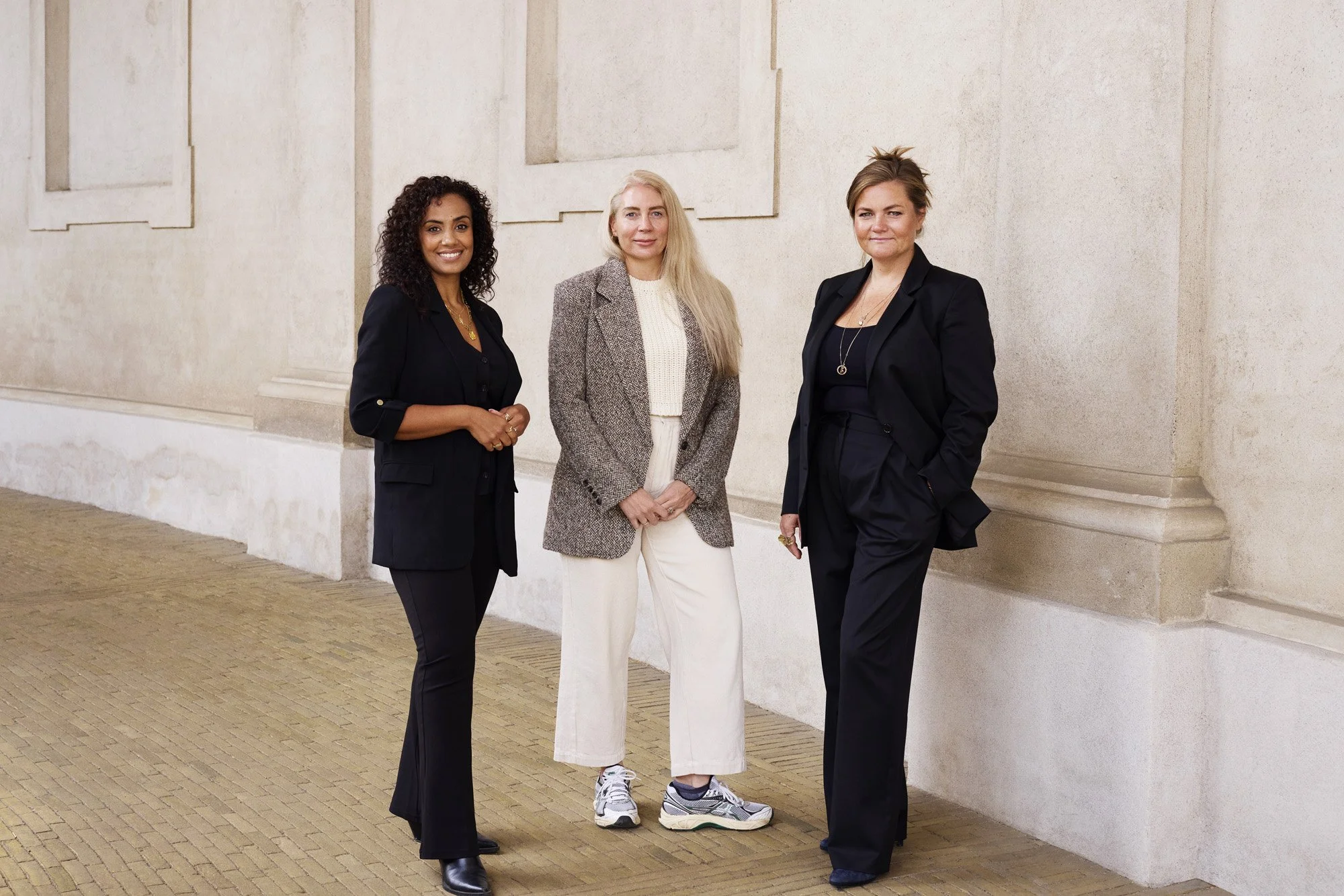 Portrait from Unconventional Ventures - Three women standing in a row against a beige wall, all wearing business attire, smiling at the camera.