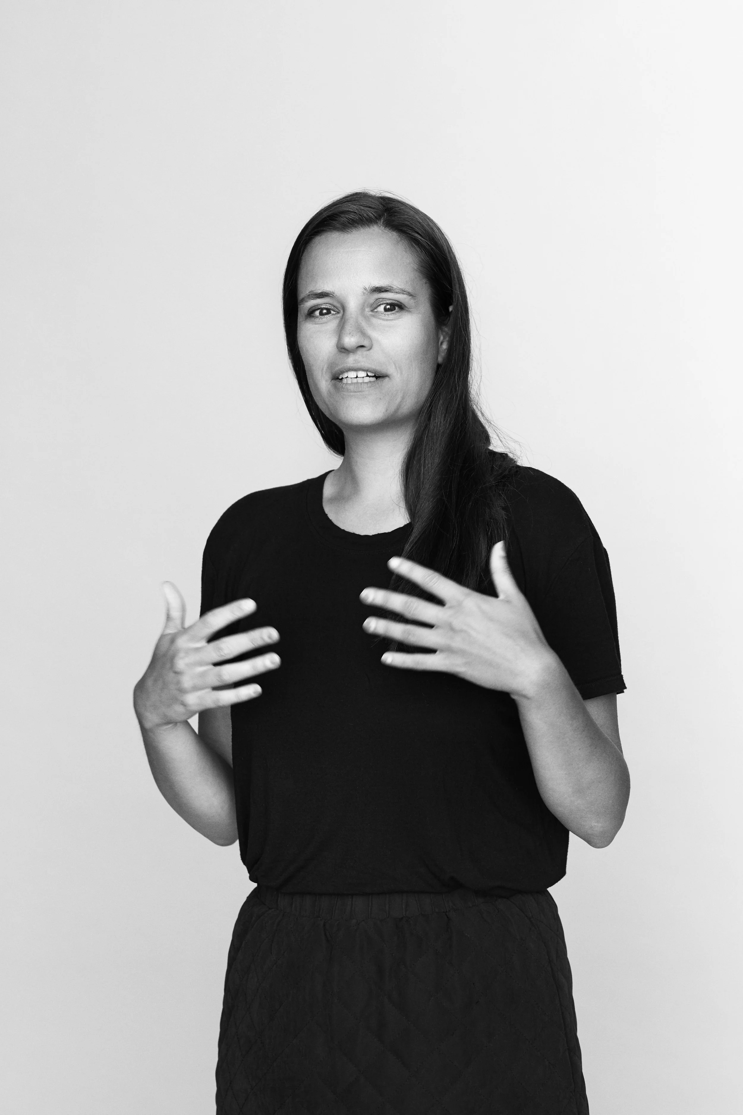 Black and white portrait of a woman with long dark hair, wearing a black T-shirt and a quilted skirt, standing against a plain background, gesturing with her hands near her chest.