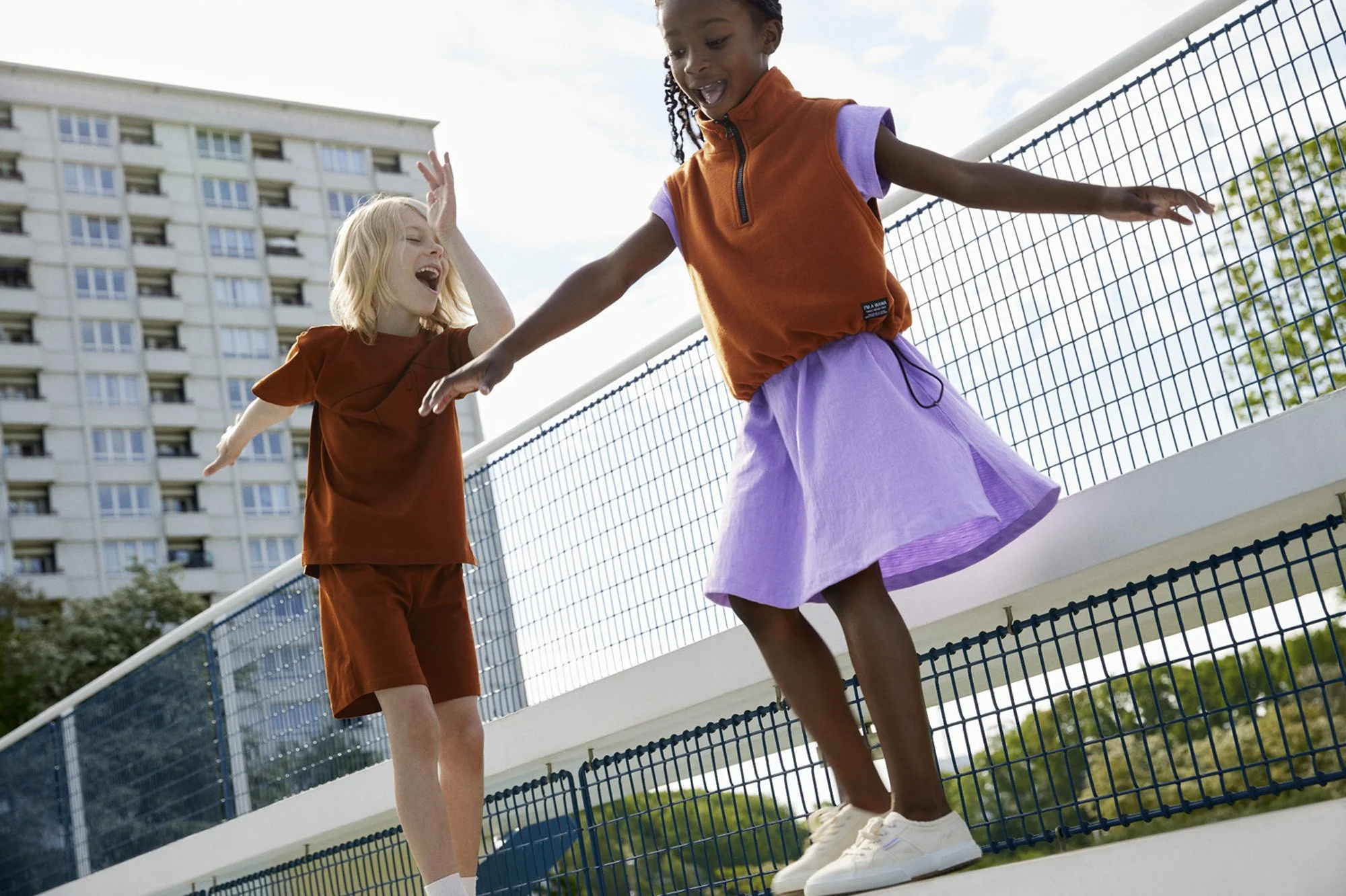WAWA CPH - Two young girls joyfully playing and balancing on a ledge outdoors near a metal fence, with a tall apartment building in the background.