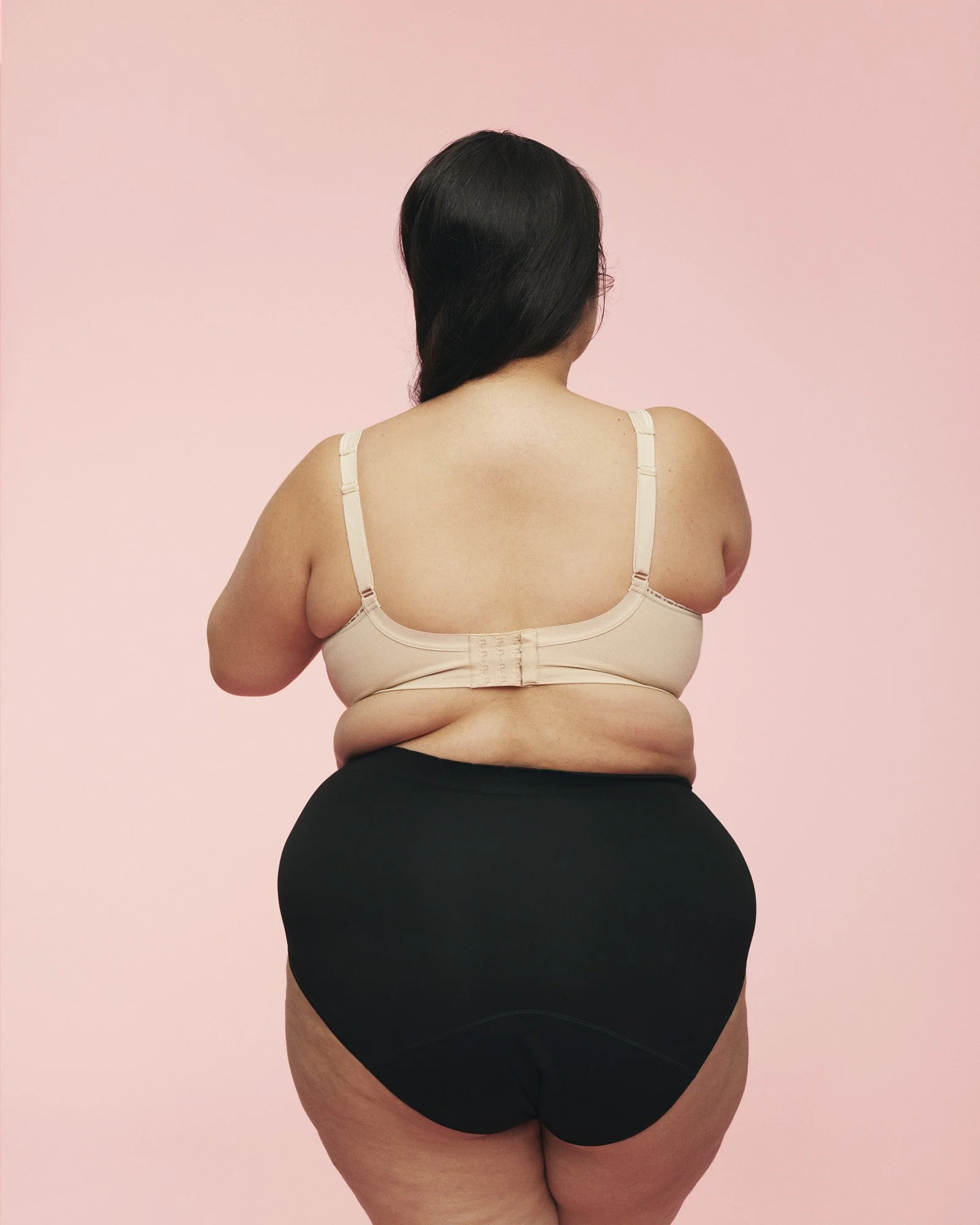 MOMKIND - Back view of a woman with long dark hair wearing a beige bra and black underwear, standing against a pink background.