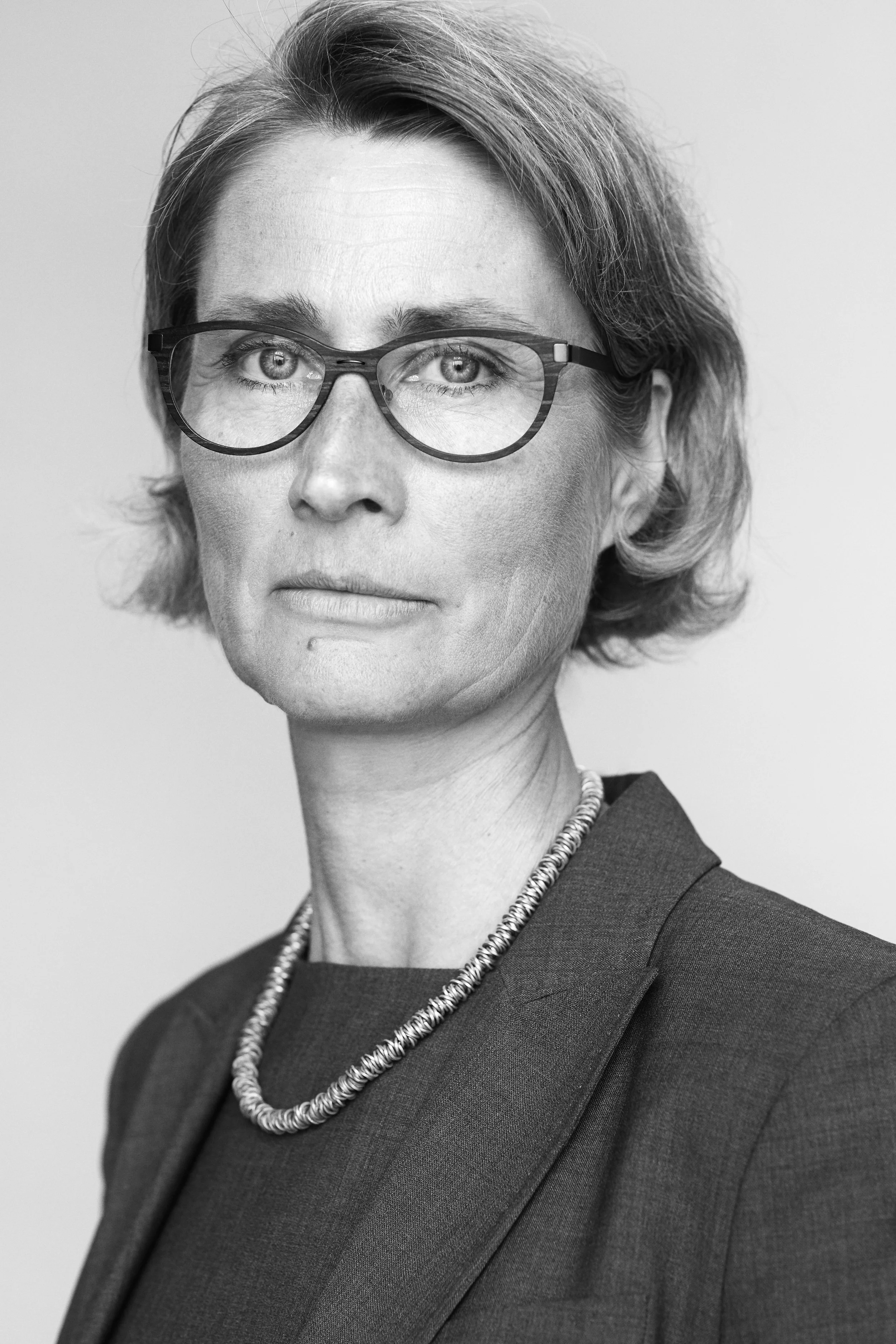 Black and white portrait of a middle-aged woman wearing glasses, a blazer, and a necklace, looking directly at the camera.