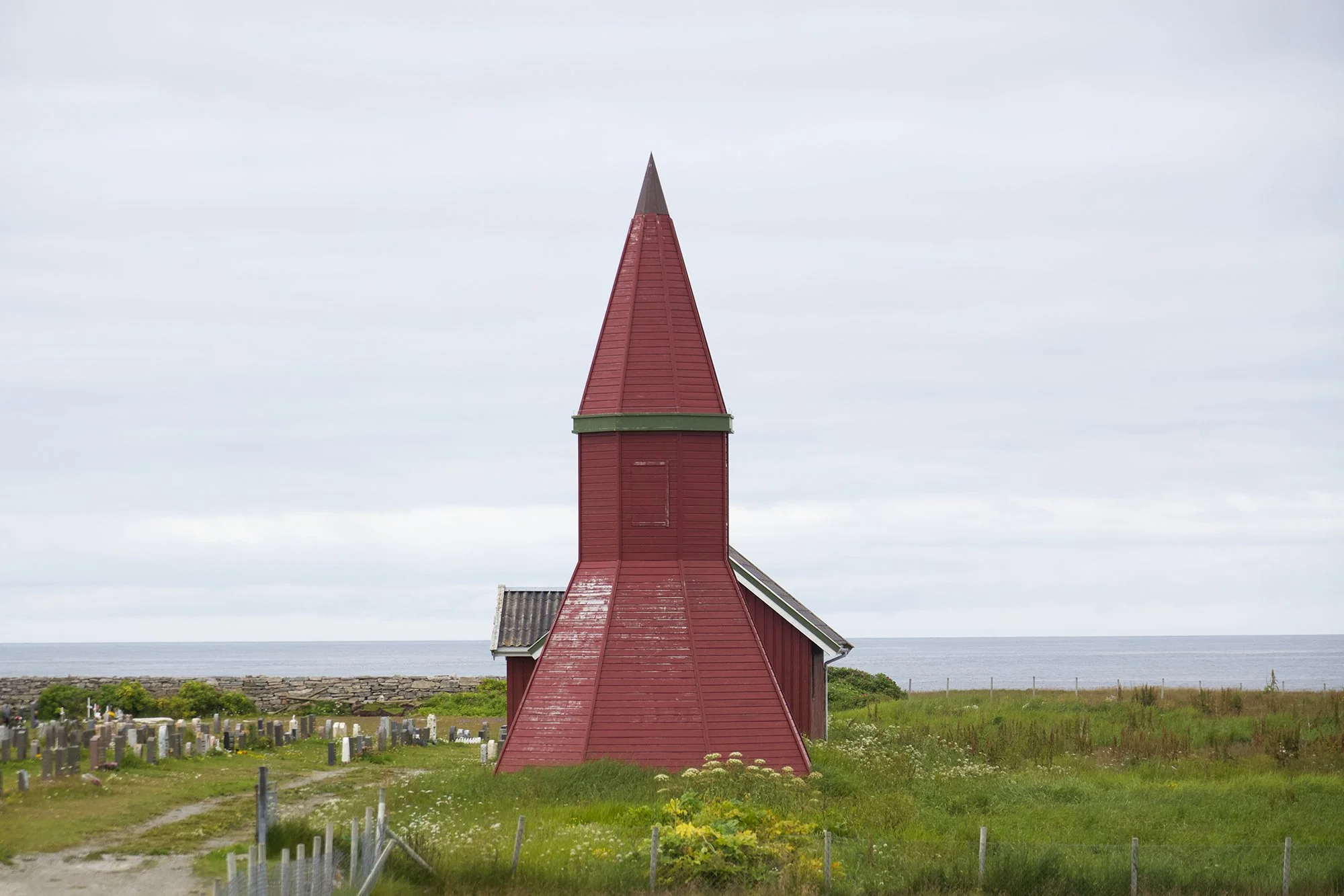 Rødt trekledd kirketårn på Røst kirke, med grønn gesims og en spiss topp dekket av metall. Malingen på tårnet er slitt flere steder, og himmelen bak er overskyet.