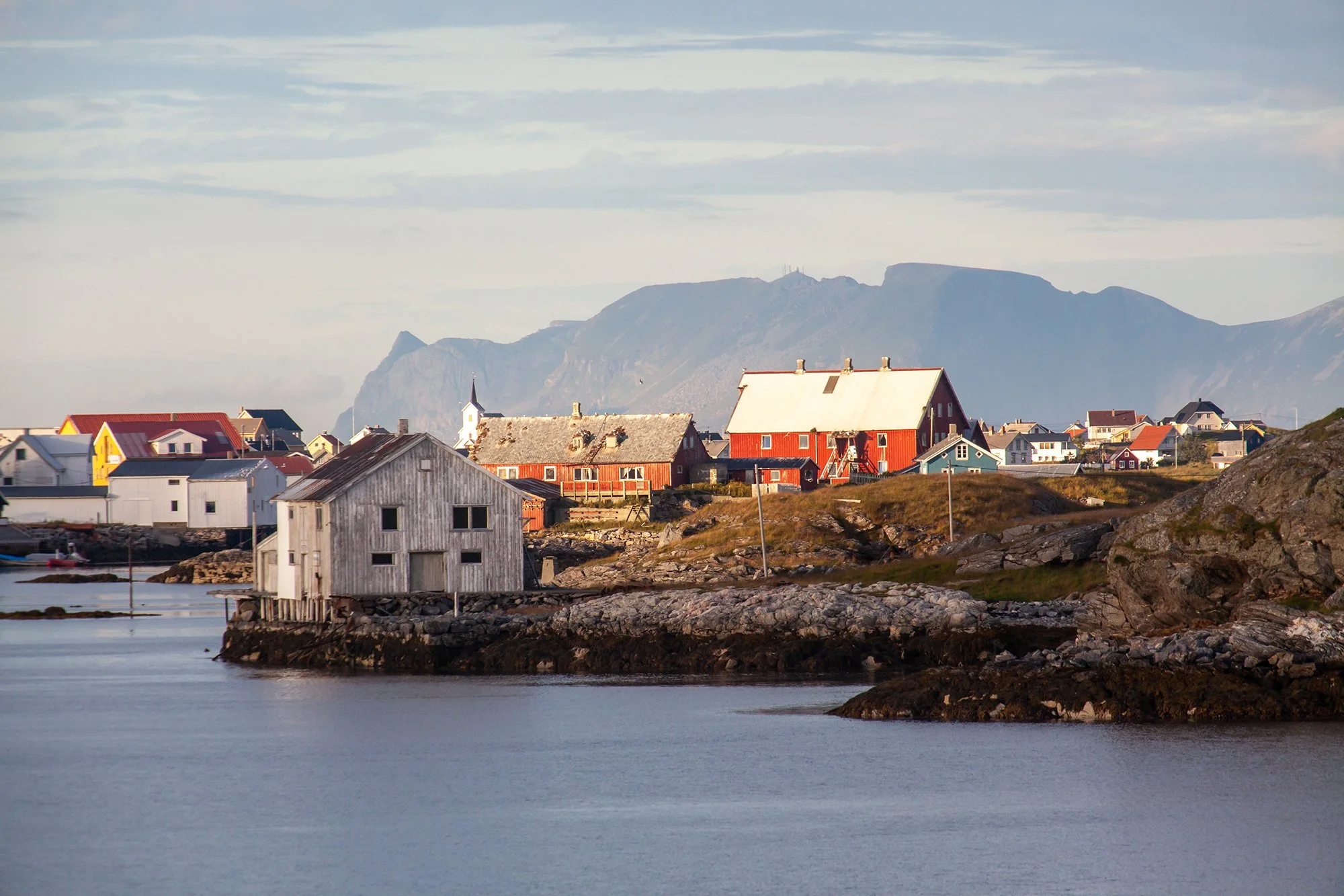 Bilde som viser bebyggelse på Røst. Et gammelt naust i forkant og fjellside i horisonten.
