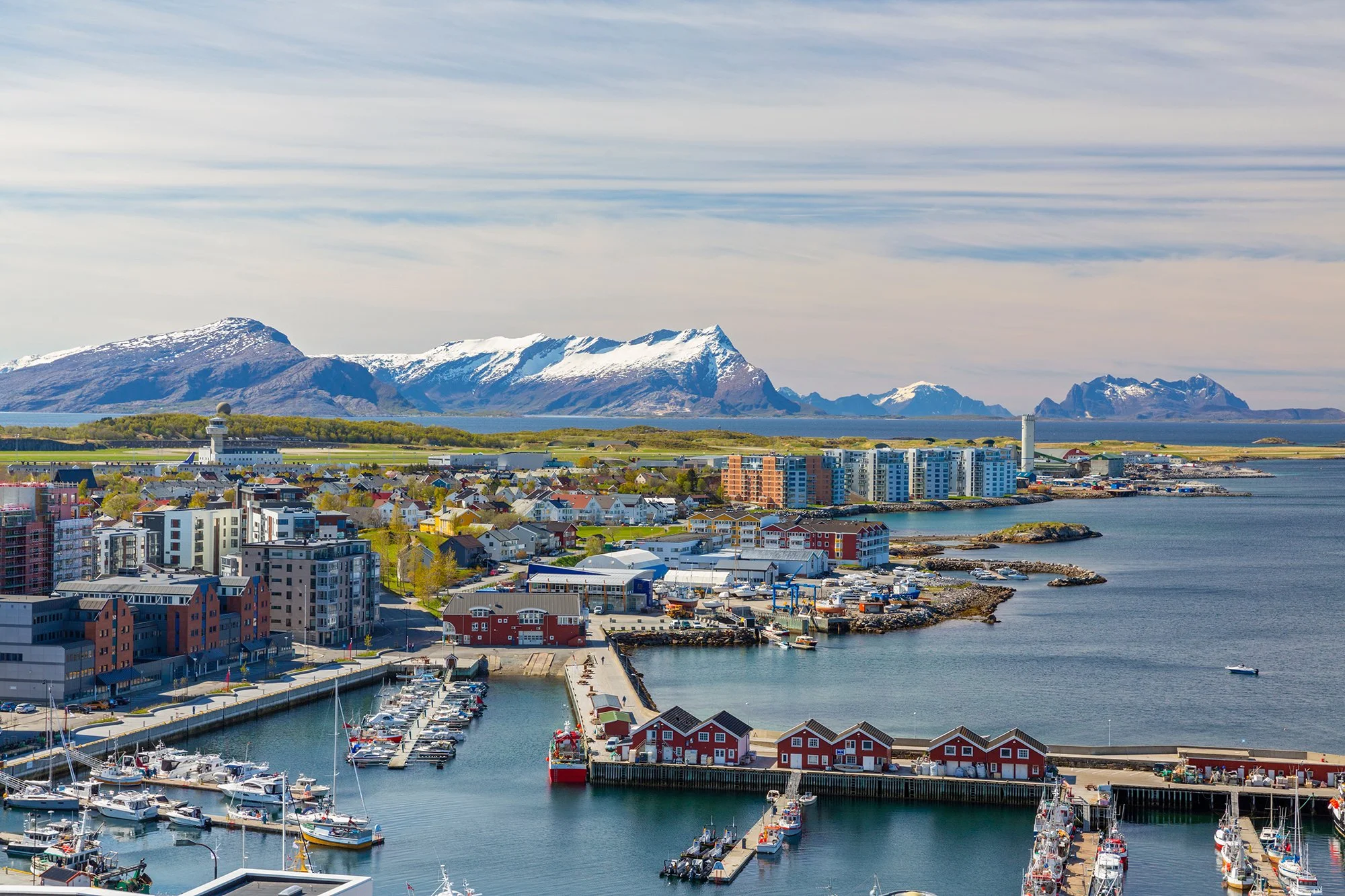 Utsiktsbilde over havnen i Bodø en tidlig vårdag. Bildet viser en mol med båter i forkant, bebyggelse i midten av bildet og fjellformasjoner bak. Det er fortsatt snø i fjellene.