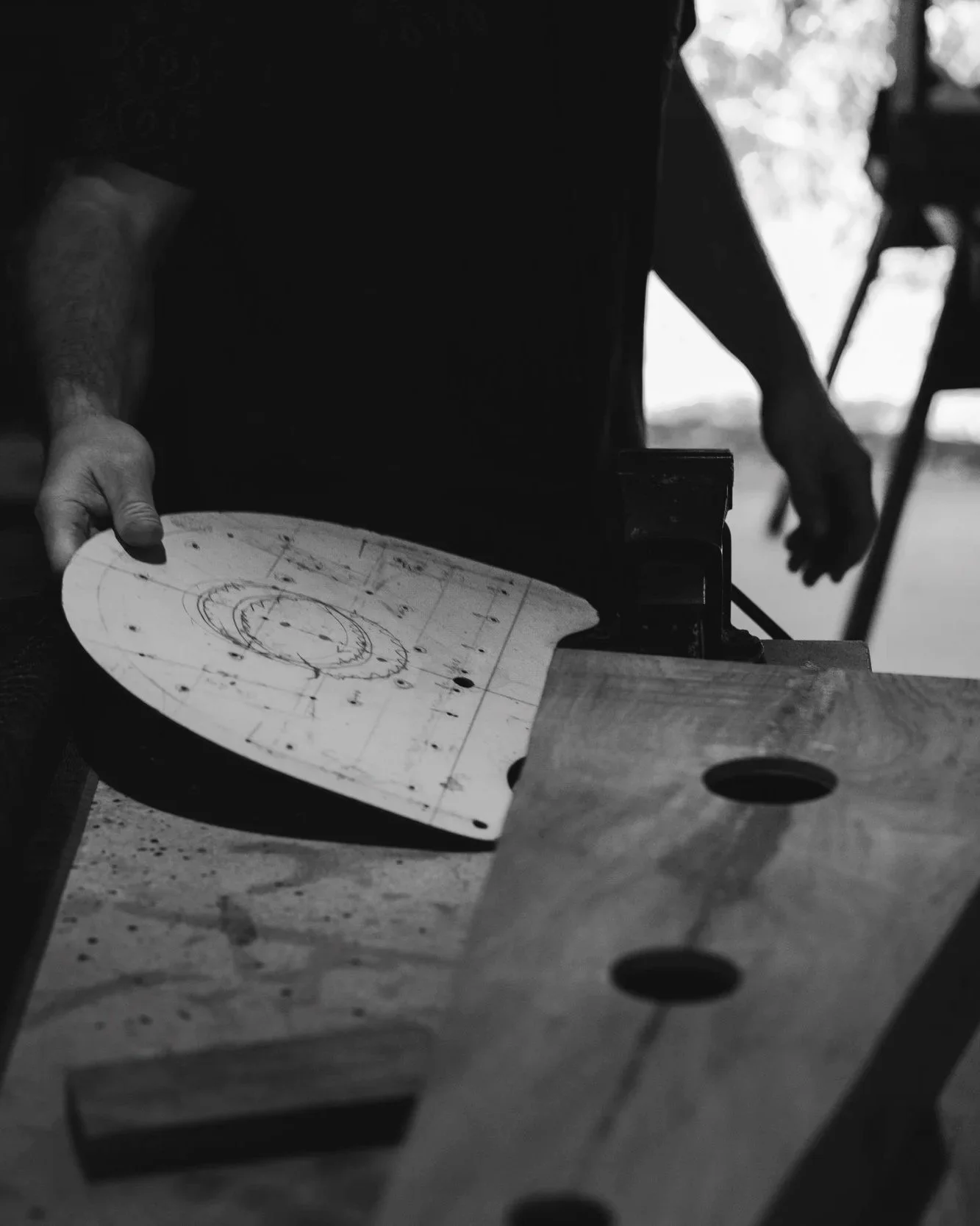Black and white photo of a person holding a round wooden board with sketches and markings, possibly in a woodworking or crafting environment, with additional woodworking tools in the background.