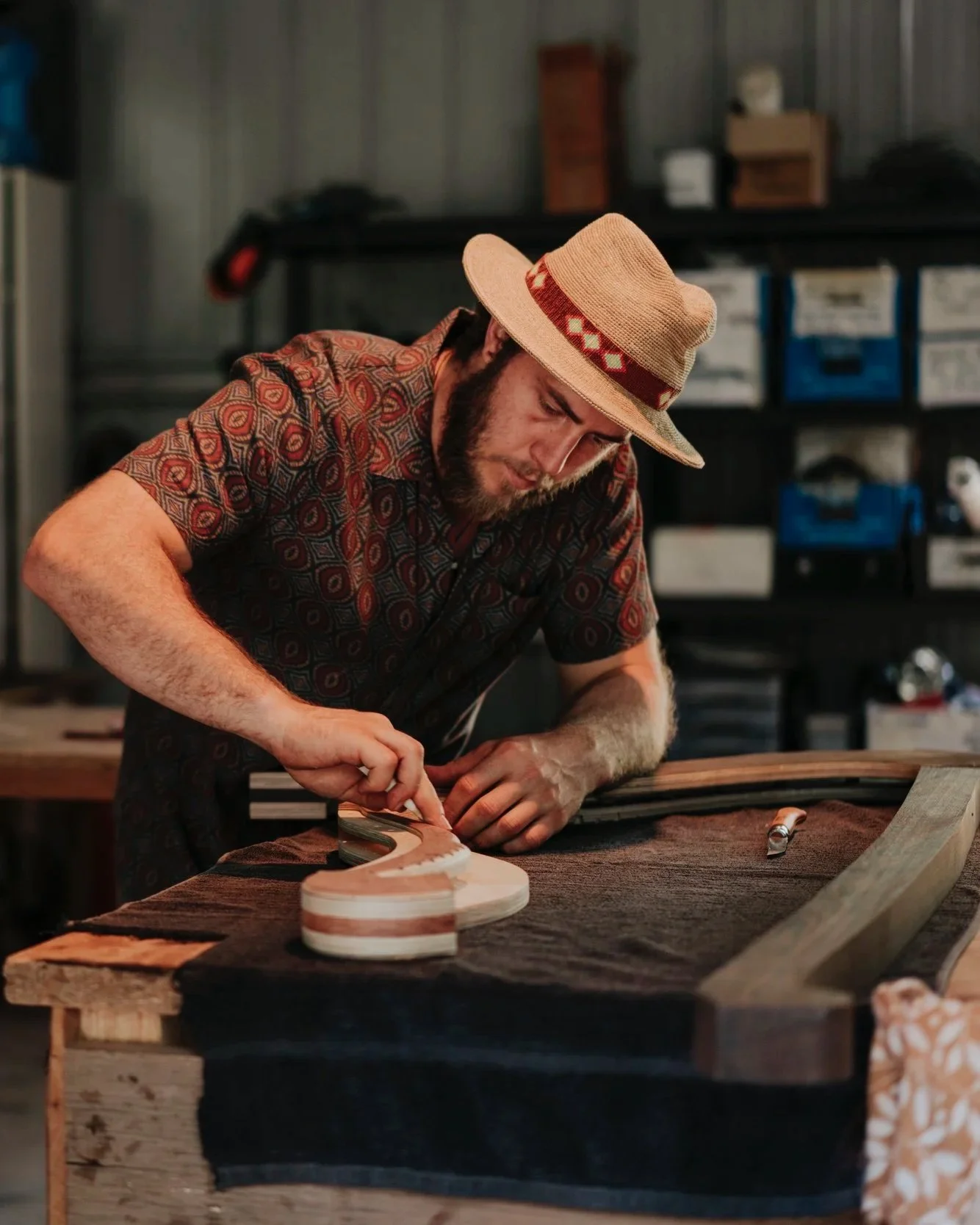 A man wearing a patterned shirt and a straw hat working on a woodworking project, shaping a piece of wood on a workbench in a workshop.