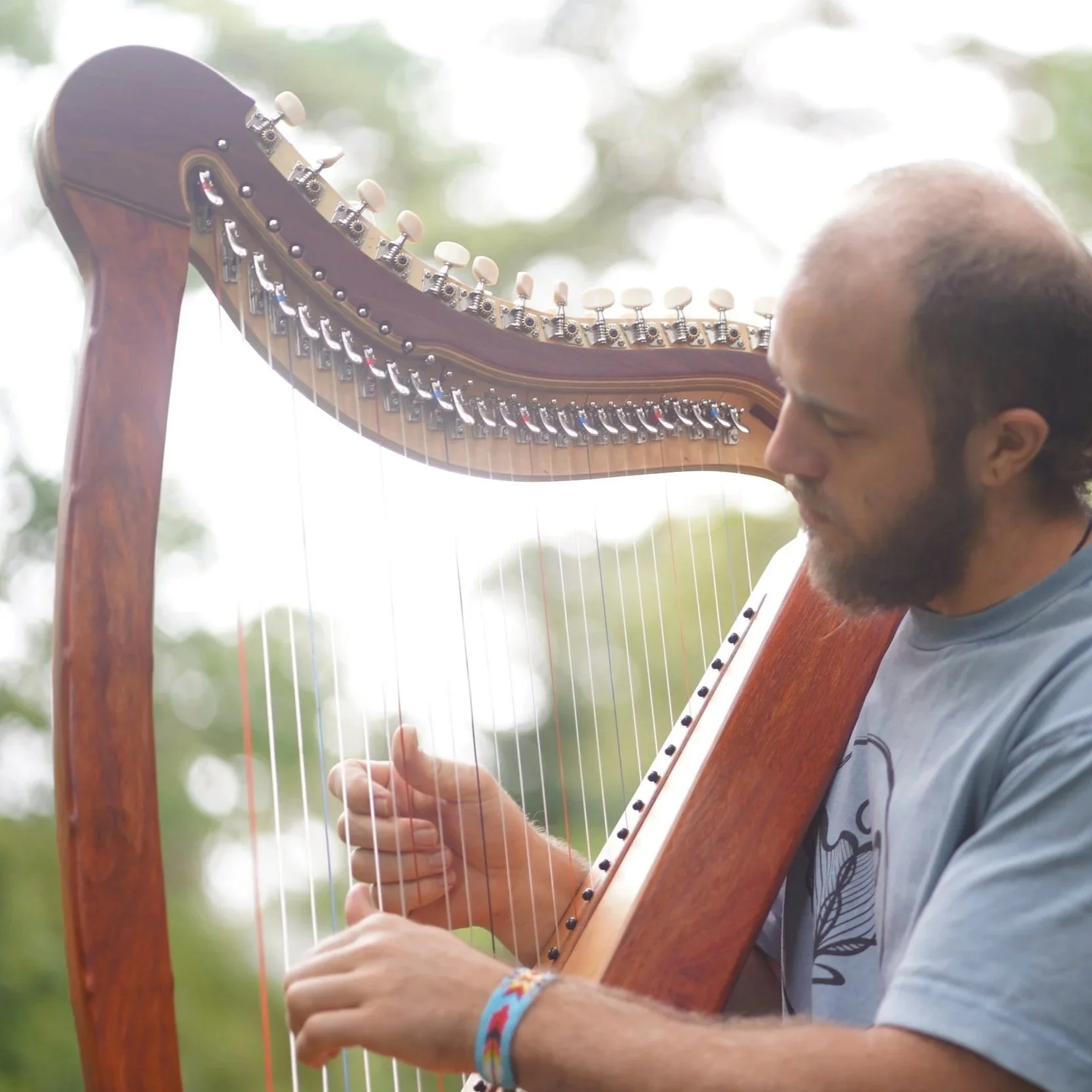 A man with a beard and a wristband playing a wooden harp outdoors, with a blurred background of trees and sky.