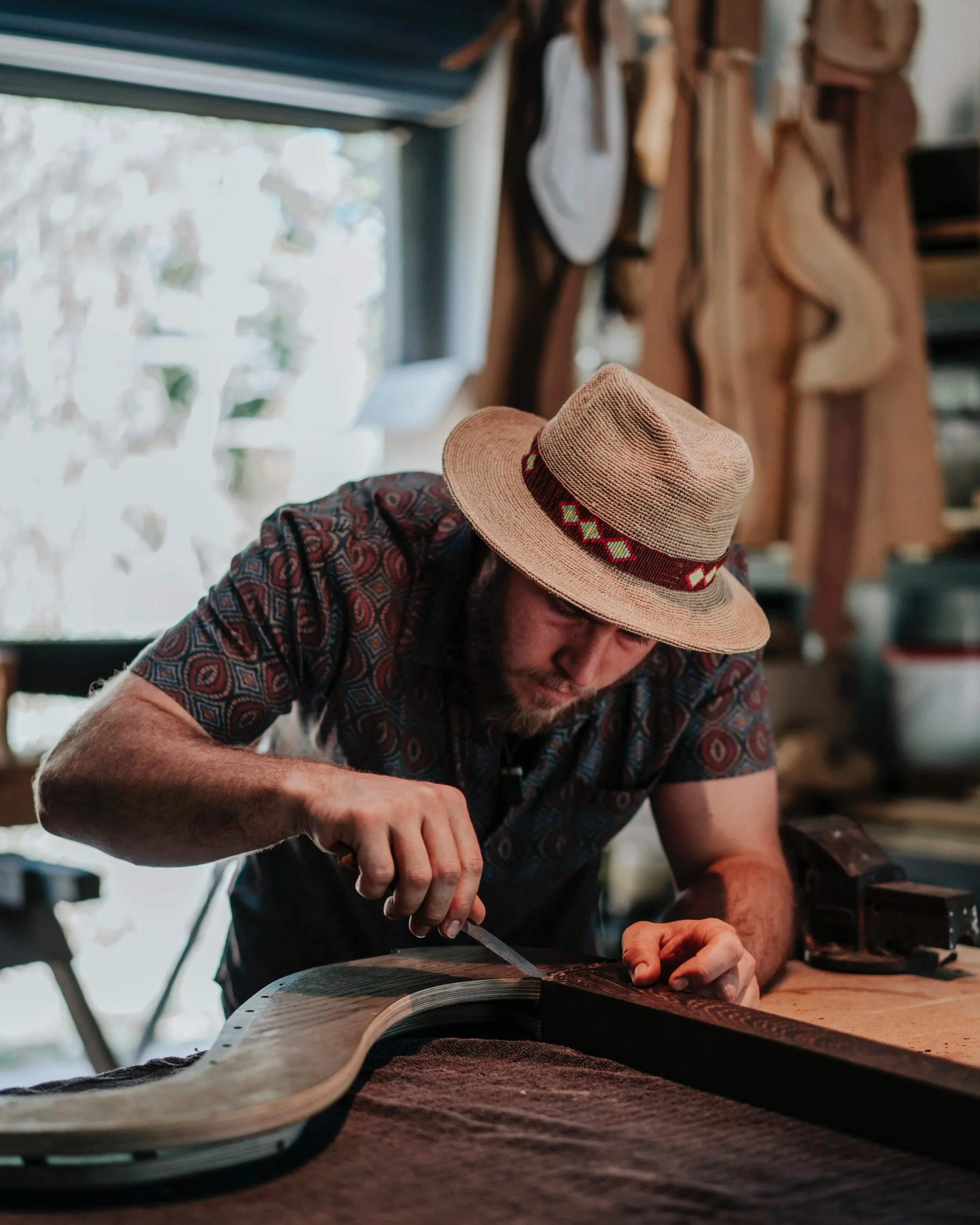 A man wearing a straw hat with a colorful band is woodworking in a workshop, using a small tool on a piece of wood.