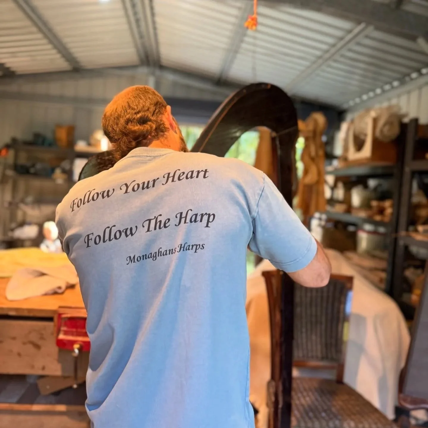 A man with brown hair, wearing a light blue t-shirt with a quote about following the heart and discovering the harp, working on a large harp inside a workshop with shelves and tools in the background.