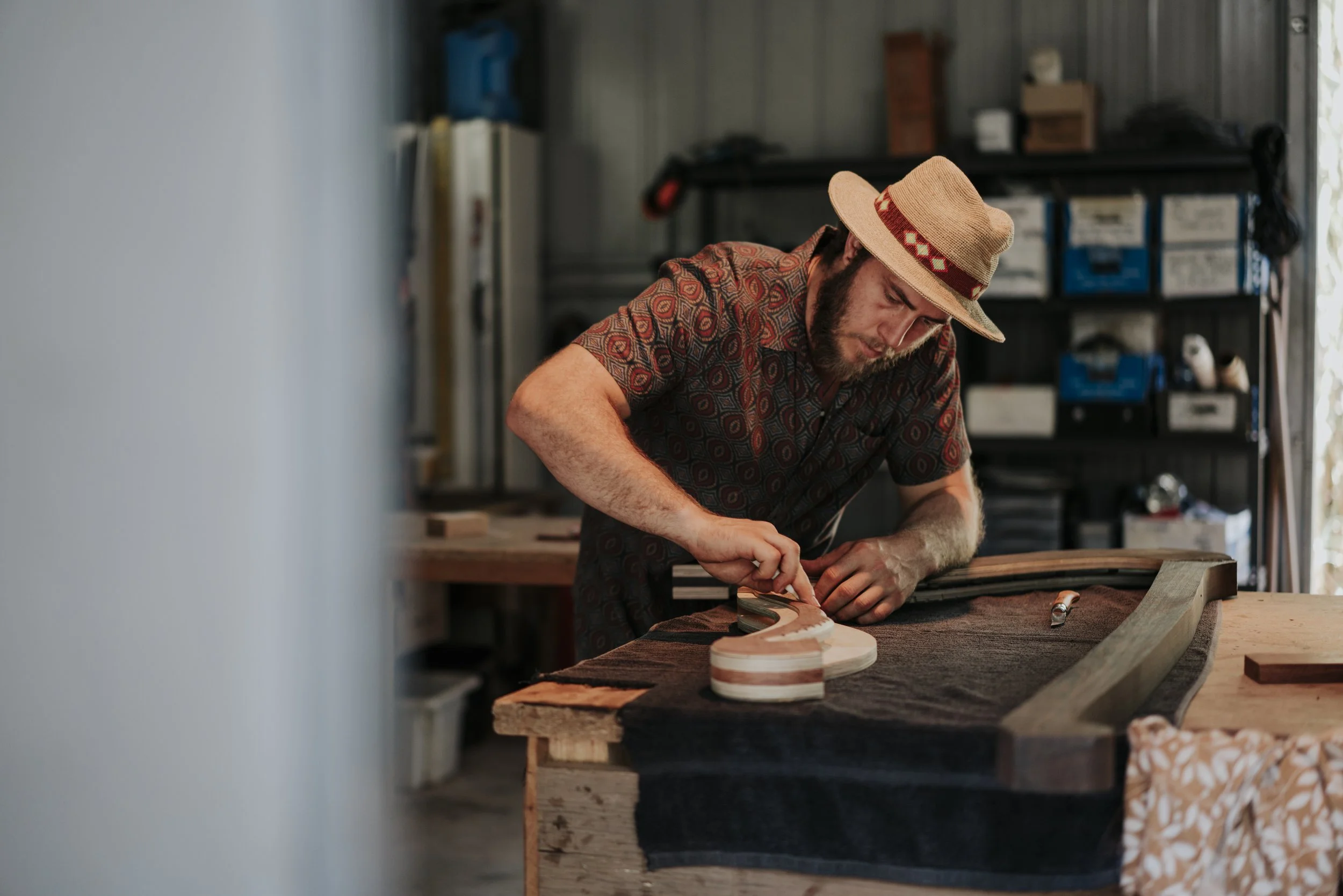 A man wearing a patterned shirt and a straw hat working on a woodworking project in a workshop, shaping a piece of wood.